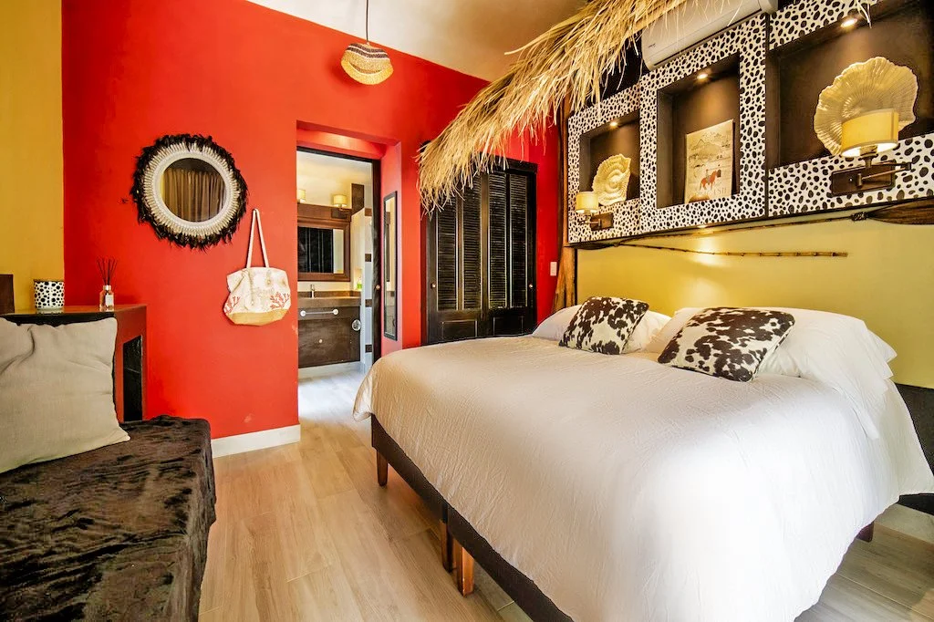 A colorful bedroom with a red wall, a large bed with animal print pillows, wall niches with shell decor, and a doorway leading to a bathroom with wooden accents.