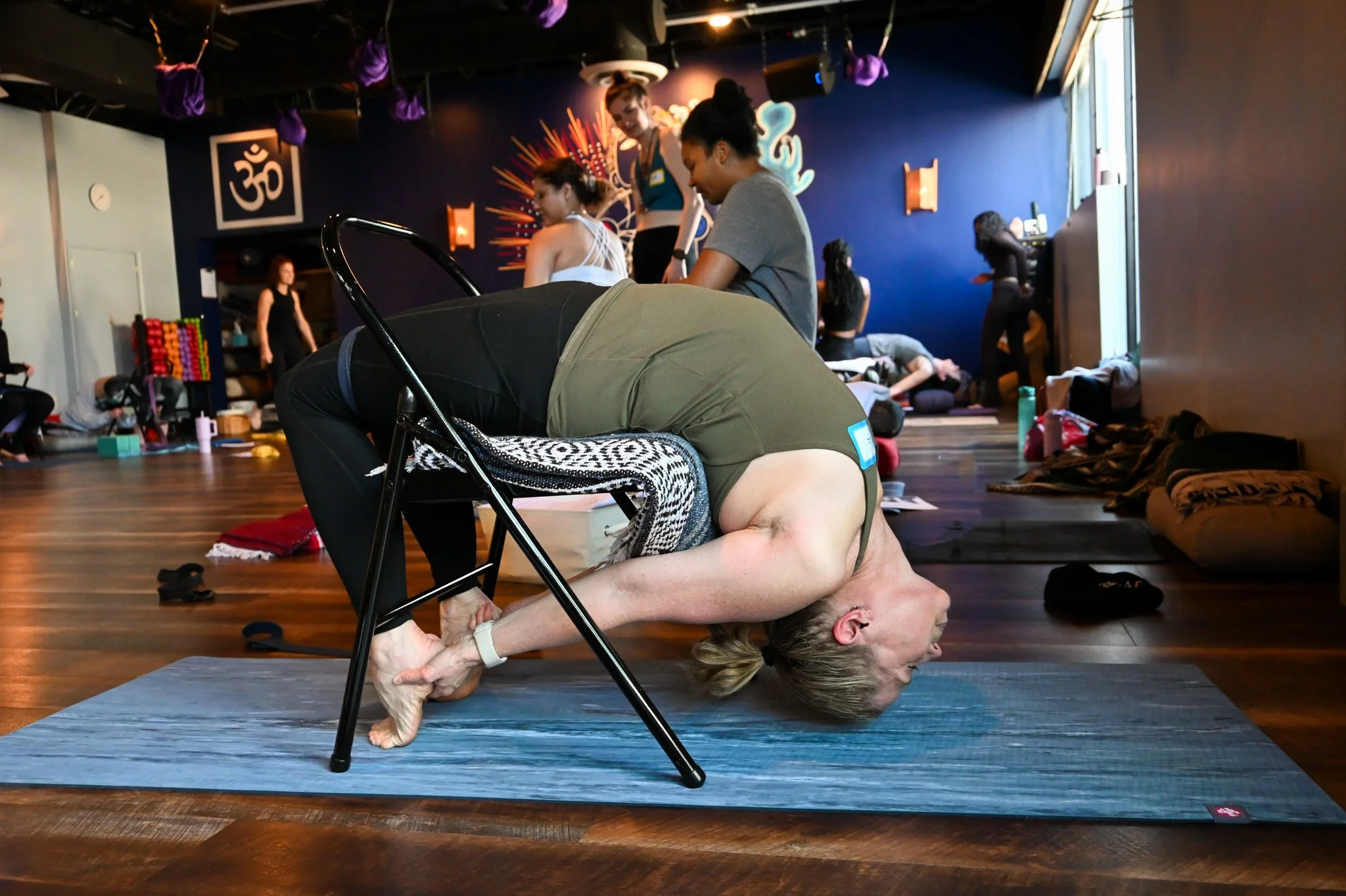 A woman practicing yoga in a child's pose with a yoga block on her back, in a studio with other people and colorful decor in the background.