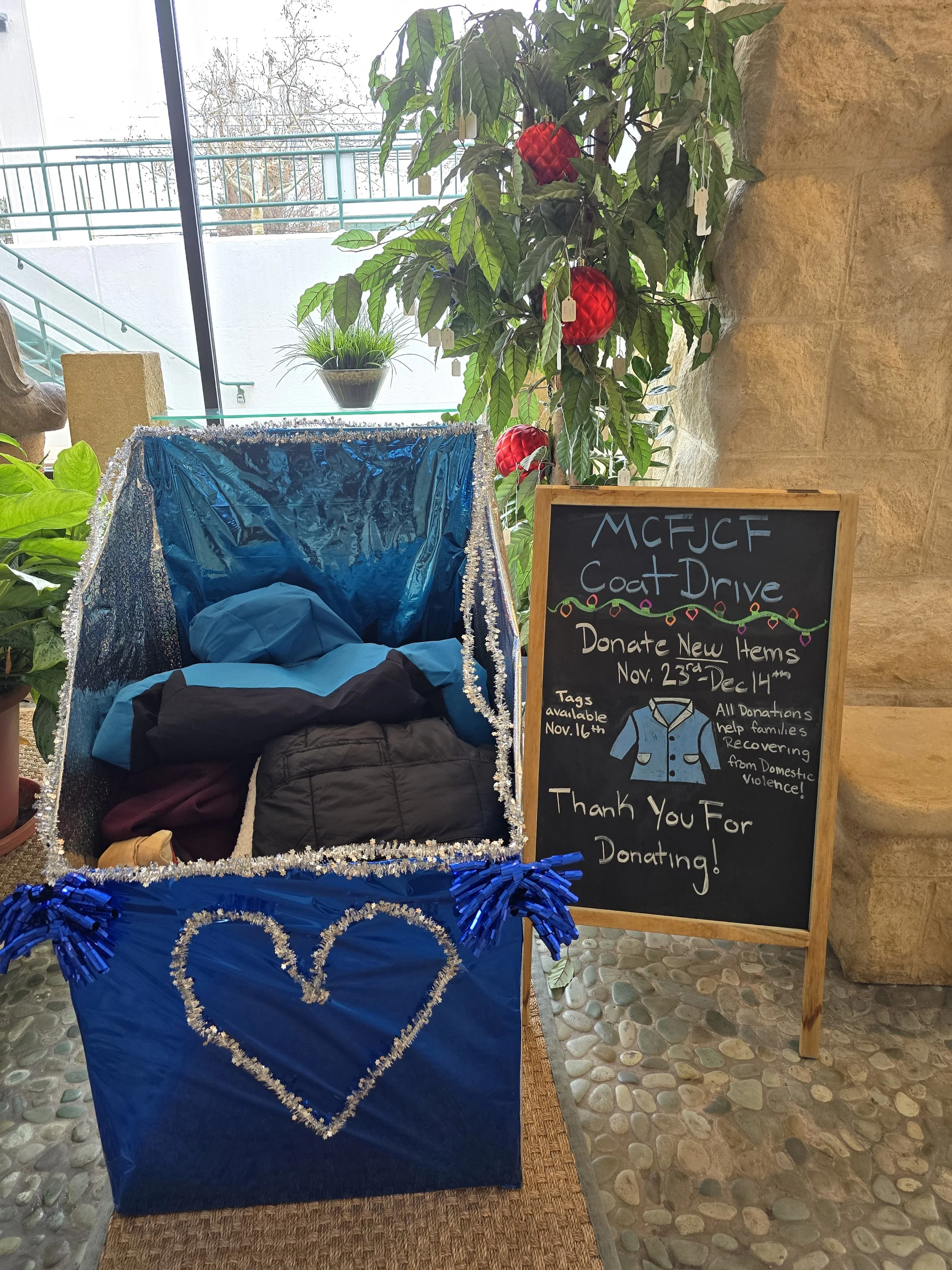 A decorated blue donation collection box for a coat drive, with a chalkboard sign next to it. The sign mentions donation dates from November 23rd to December 14th, with tags available starting November 16th. The coat drive benefits families recovering from domestic violence.