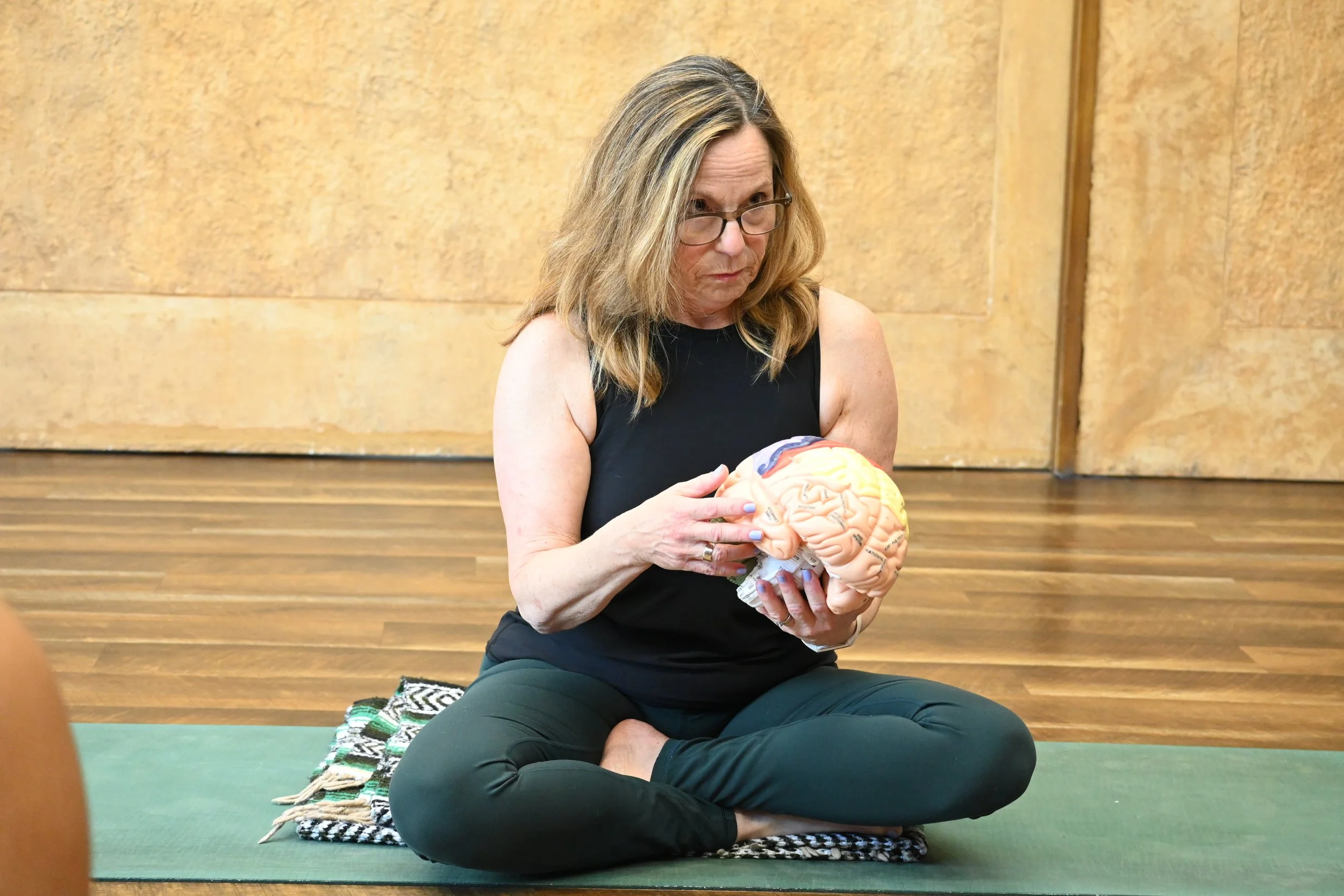A woman with glasses and shoulder-length blonde hair, wearing a black sleeveless top, sitting cross-legged on a green yoga mat, holding a detailed model of a human brain while participating in a class or workshop in a room with a wooden floor and beige wall.