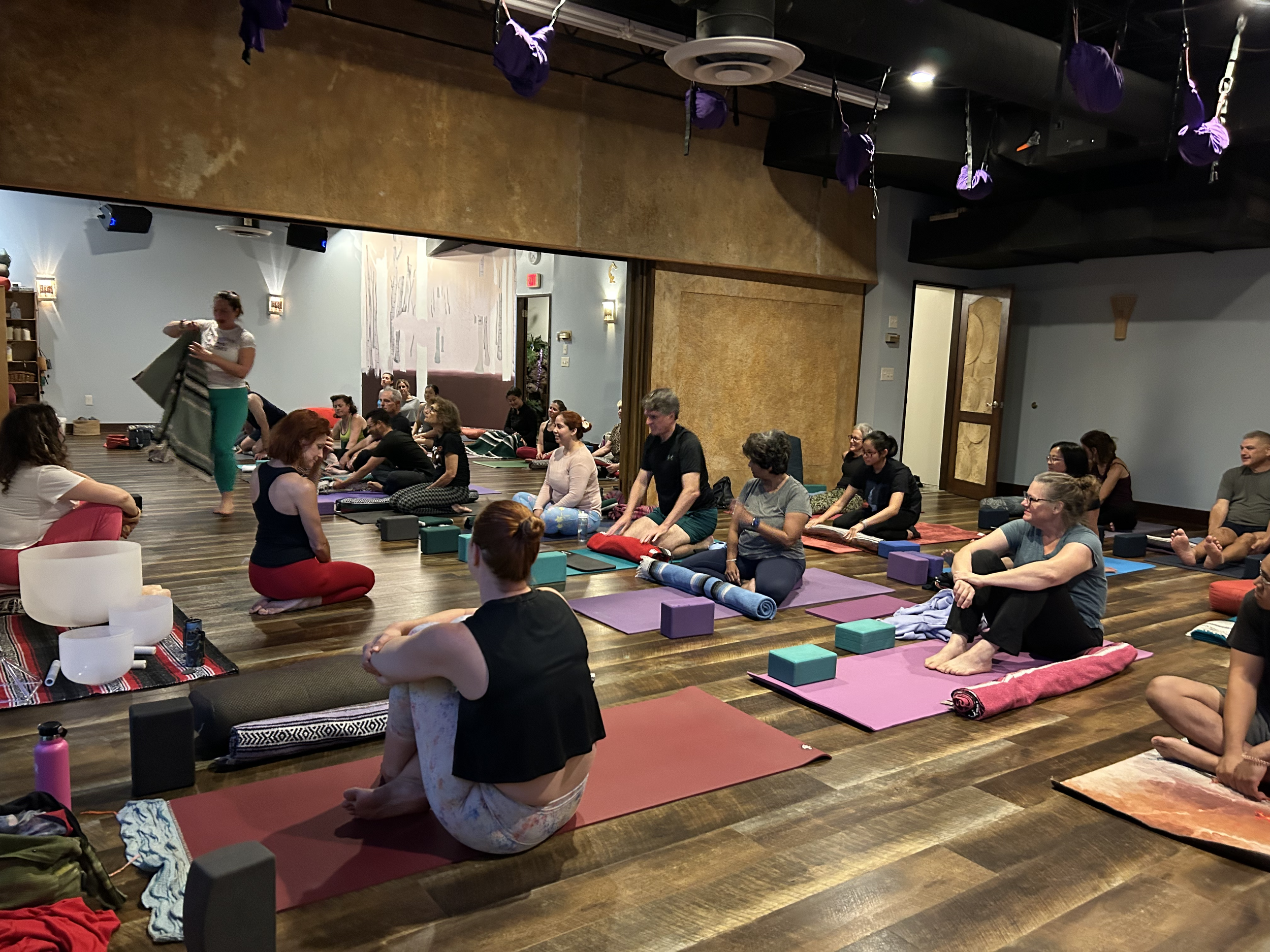 A group of people participating in a yoga or meditation class in a room with wooden floors and purple hanging hammocks or swings from the ceiling. They are seated on yoga mats in various colors, some with blocks and bags nearby, and appear to be engaged in a relaxed, mindful activity.