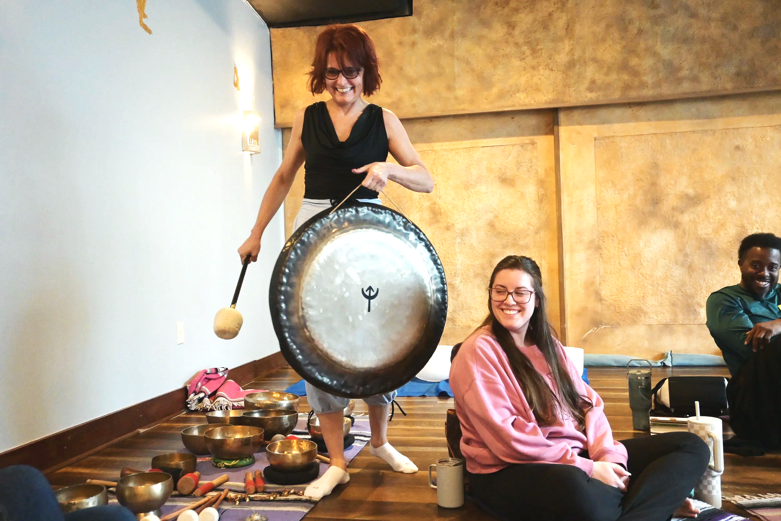 A woman with red hair and glasses smiling as she strikes a gong surrounded by various meditation bowls and instruments on the floor. Three other people, two women and a man, are sitting on the floor nearby, smiling and relaxing.