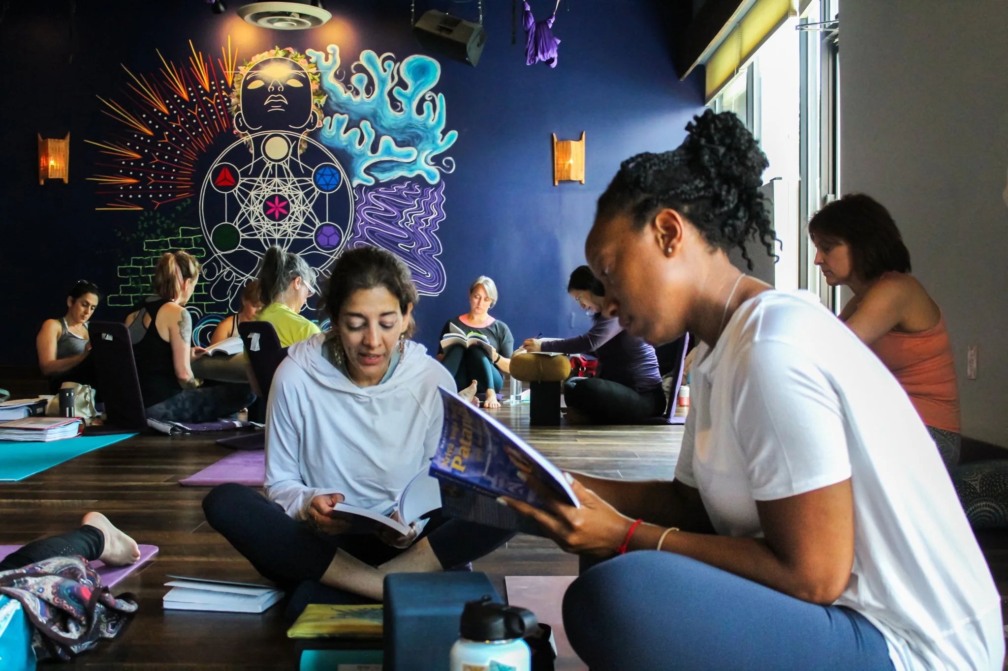 A group of women sitting on the floor in a meditation or yoga class, with one woman reading a magazine. The room has a blue wall with colorful murals, including a large face, abstract shapes, and geometric patterns, along with wall-mounted lights and a large window.