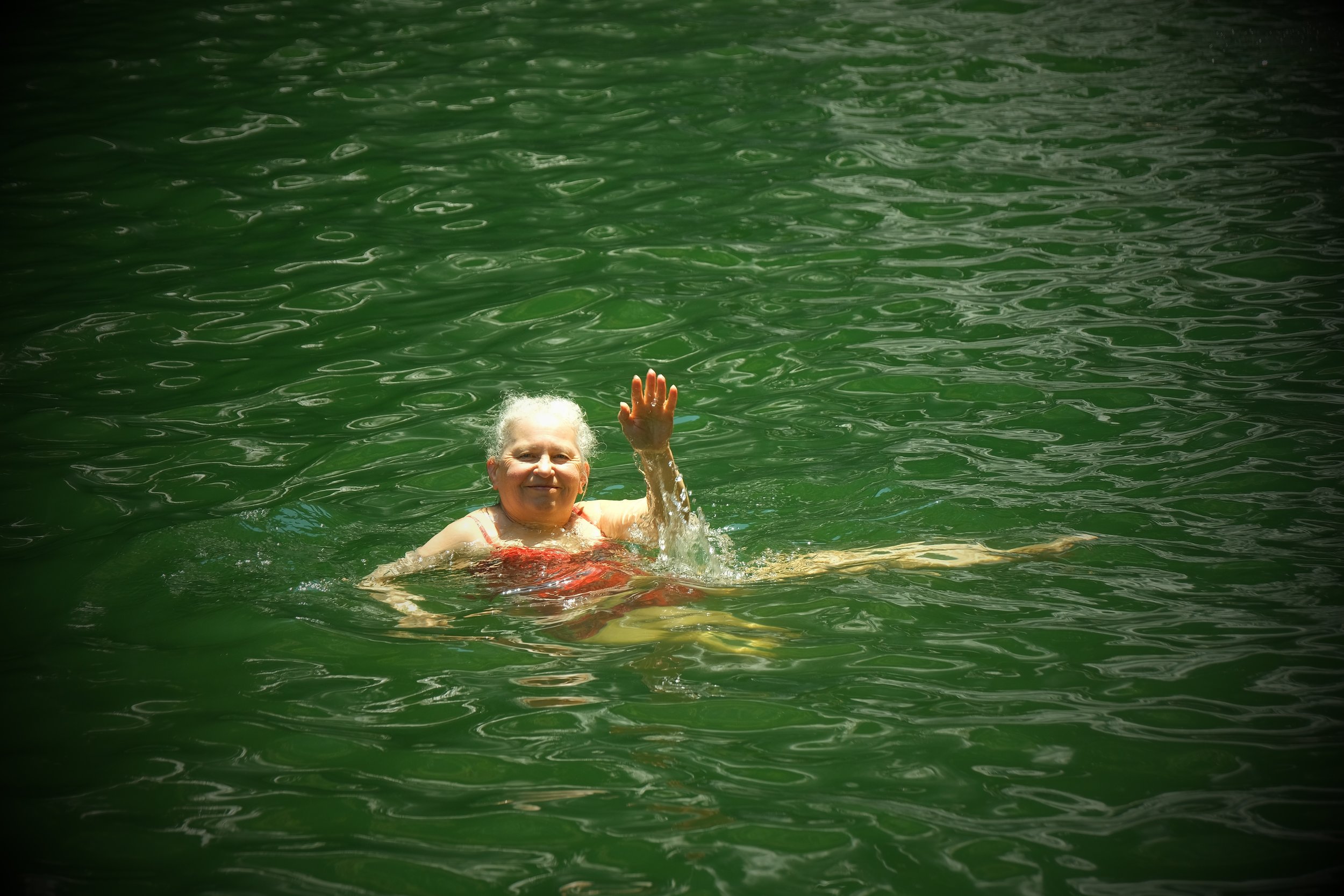 An older woman with gray hair swimming in a body of green water, smiling and waving.