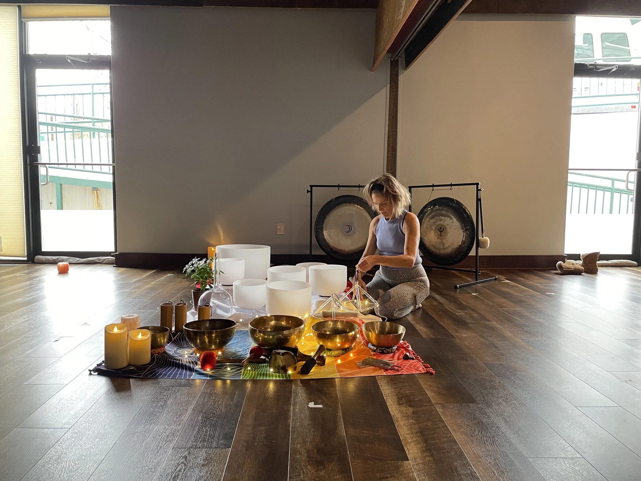 A woman participating in a sound healing or meditation session indoors with large gongs in the background. She is sitting on the floor among illuminated candles, bowls, and decorative objects placed on a colorful cloth. The room has large windows wit
