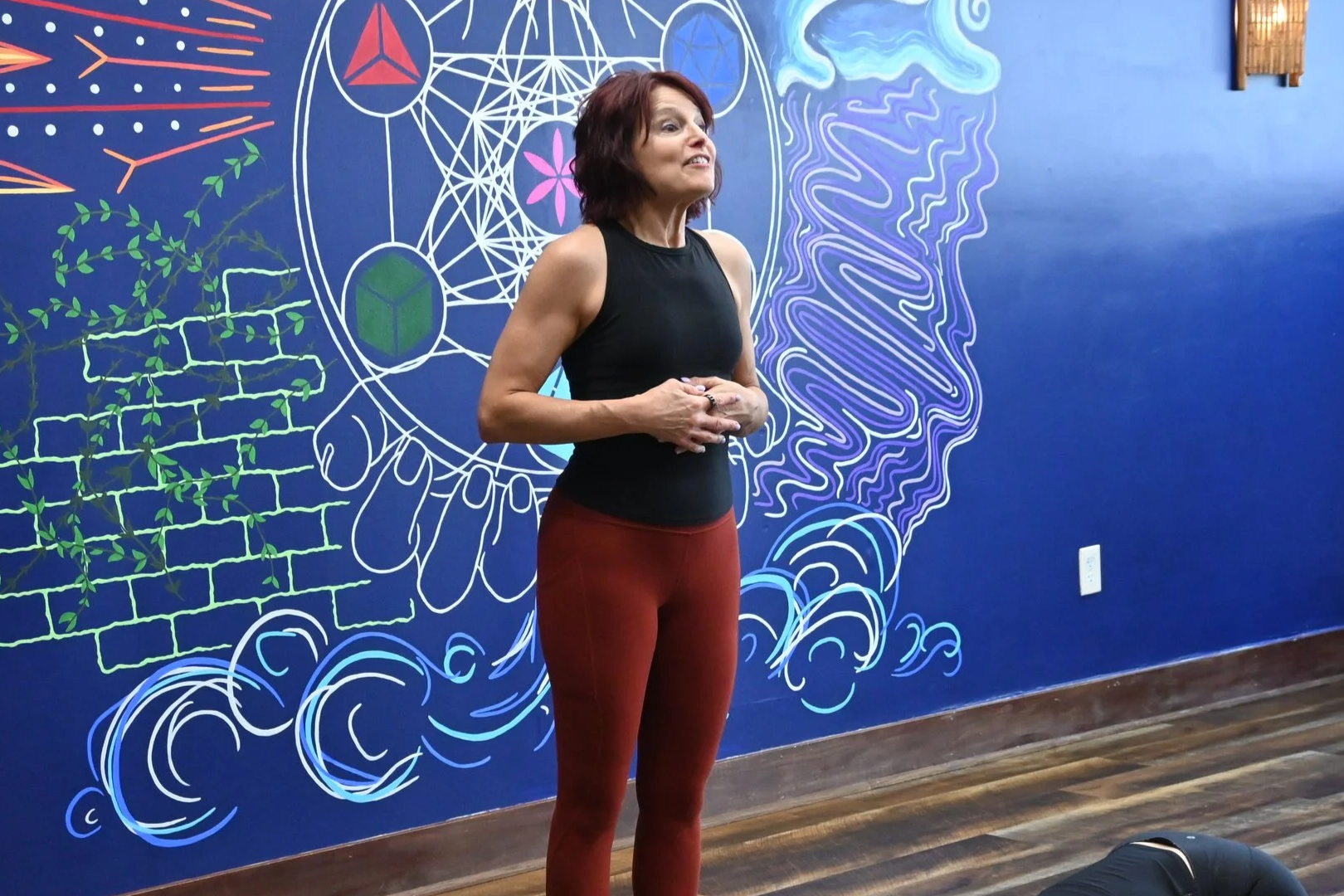A woman with short dark hair wearing a black sleeveless top and red leggings standing in front of a colorful mural with abstract shapes, a brick pattern, and a profile of a face on a blue wall.