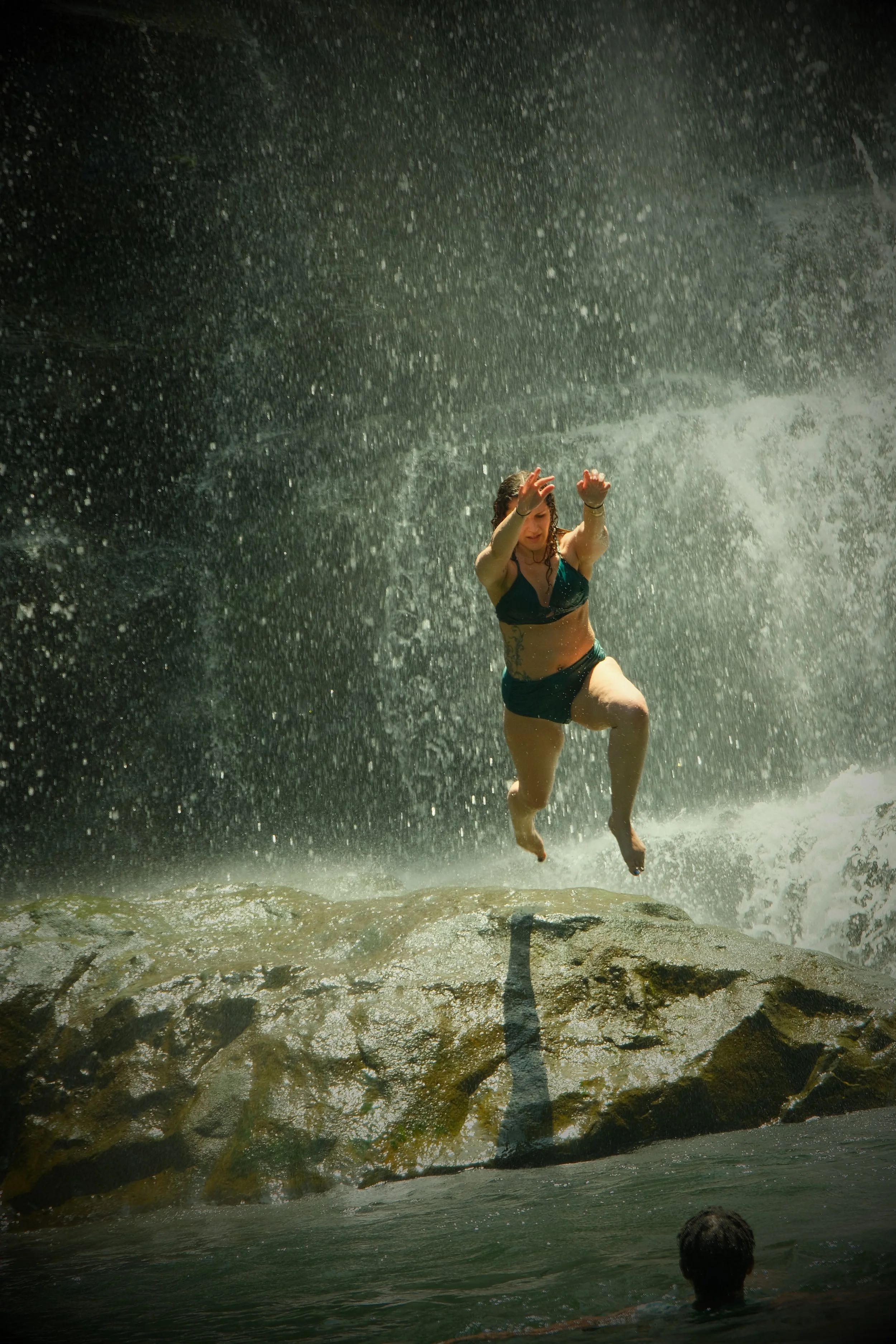 Person in a bikini jumping in front of a waterfall, with another person in the water watching.