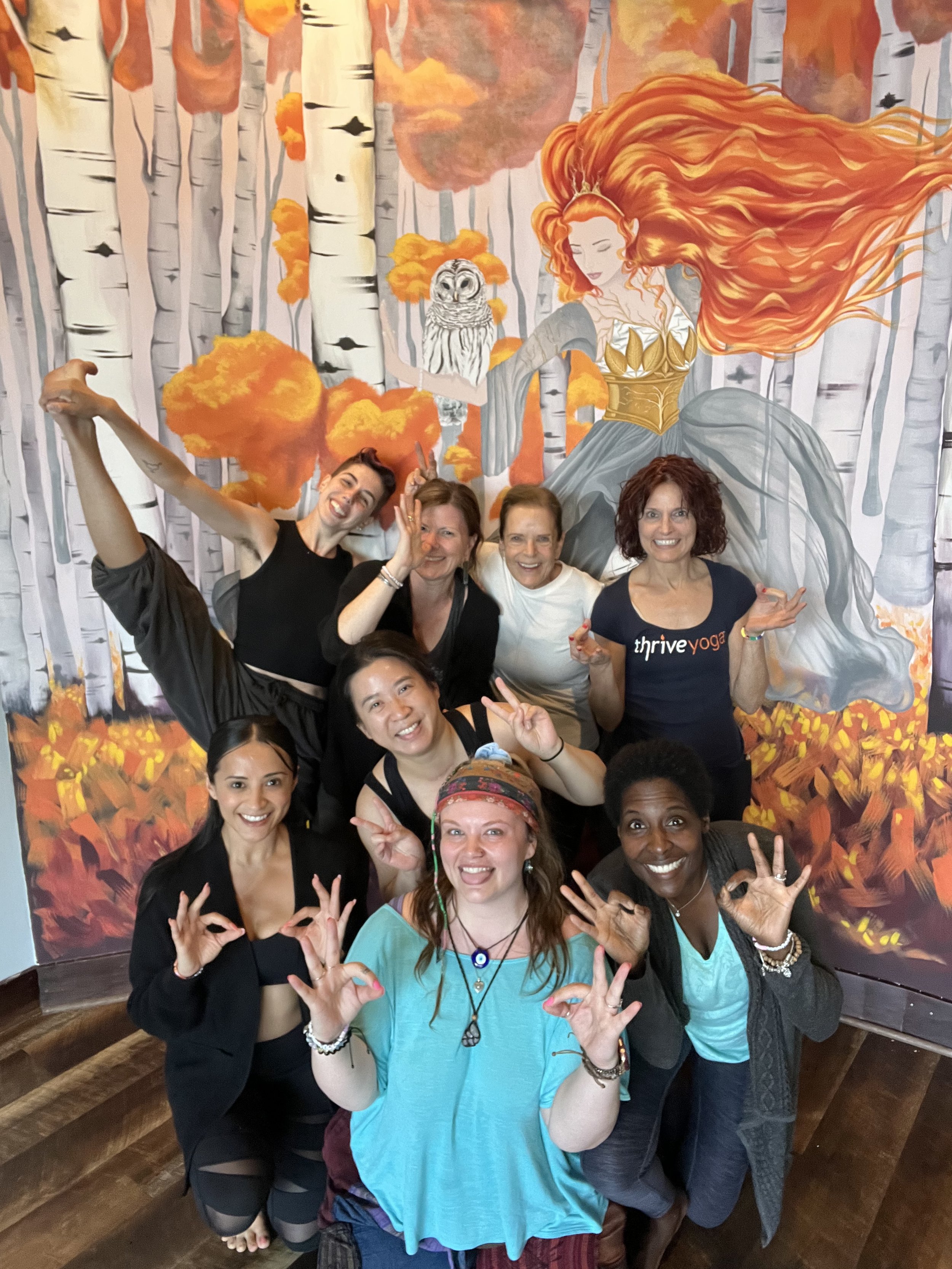 Group of smiling women posing happily in front of a mural of a fairy tale scene with a woman with flowing red hair and a snowy owl, surrounded by trees with autumn-colored leaves.