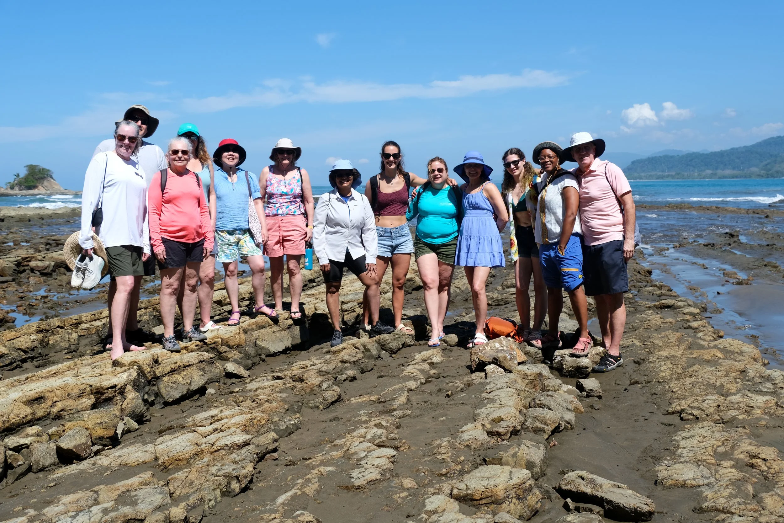 A large group of people standing together on a rocky beach, smiling at the camera with the ocean and a hilly coastline in the background on a sunny day.