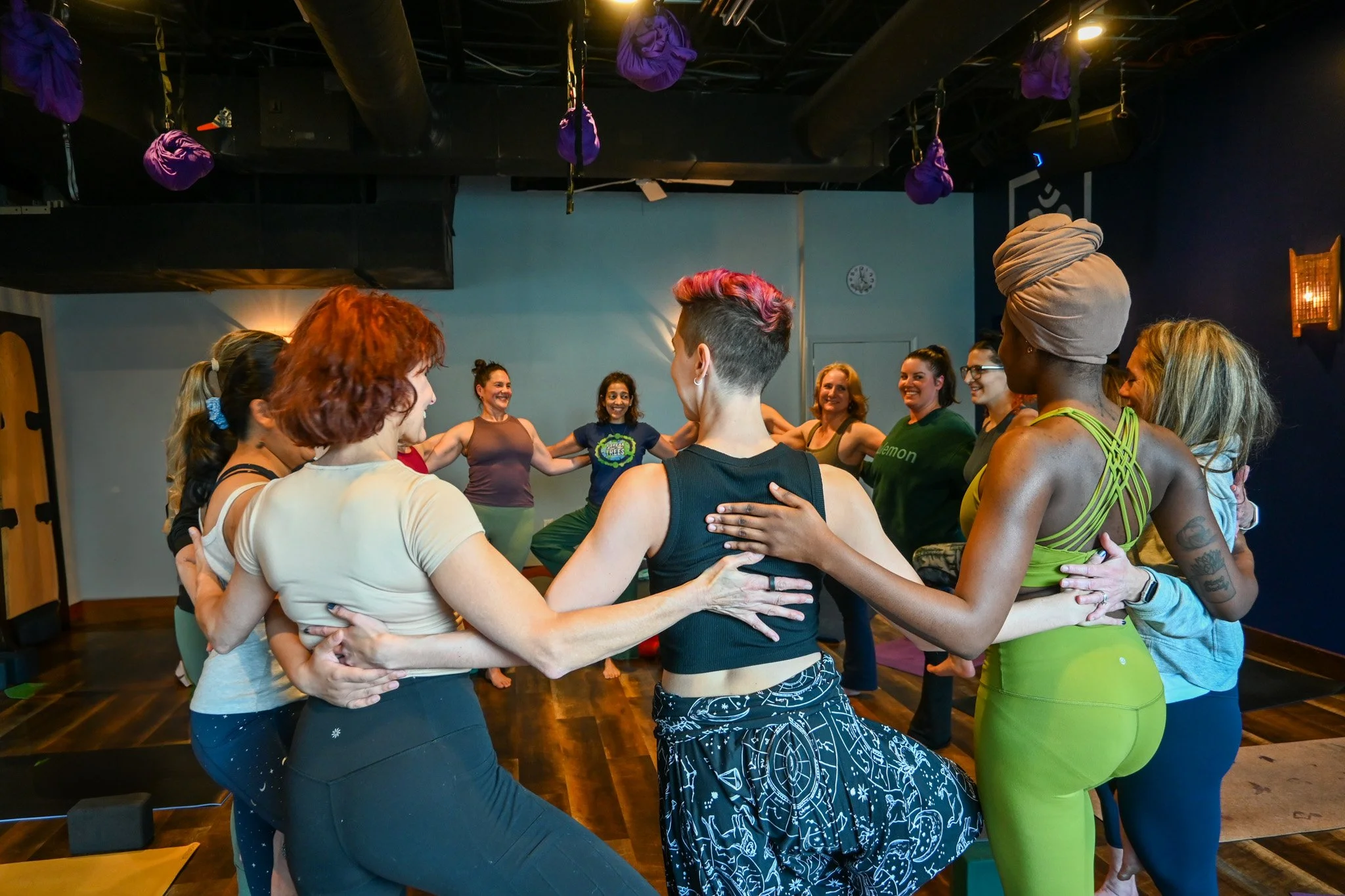 Group of women in a circle practicing partner yoga or a similar activity in a fitness studio with purple aerial yoga hammocks hanging from the ceiling. Thrive Yoga