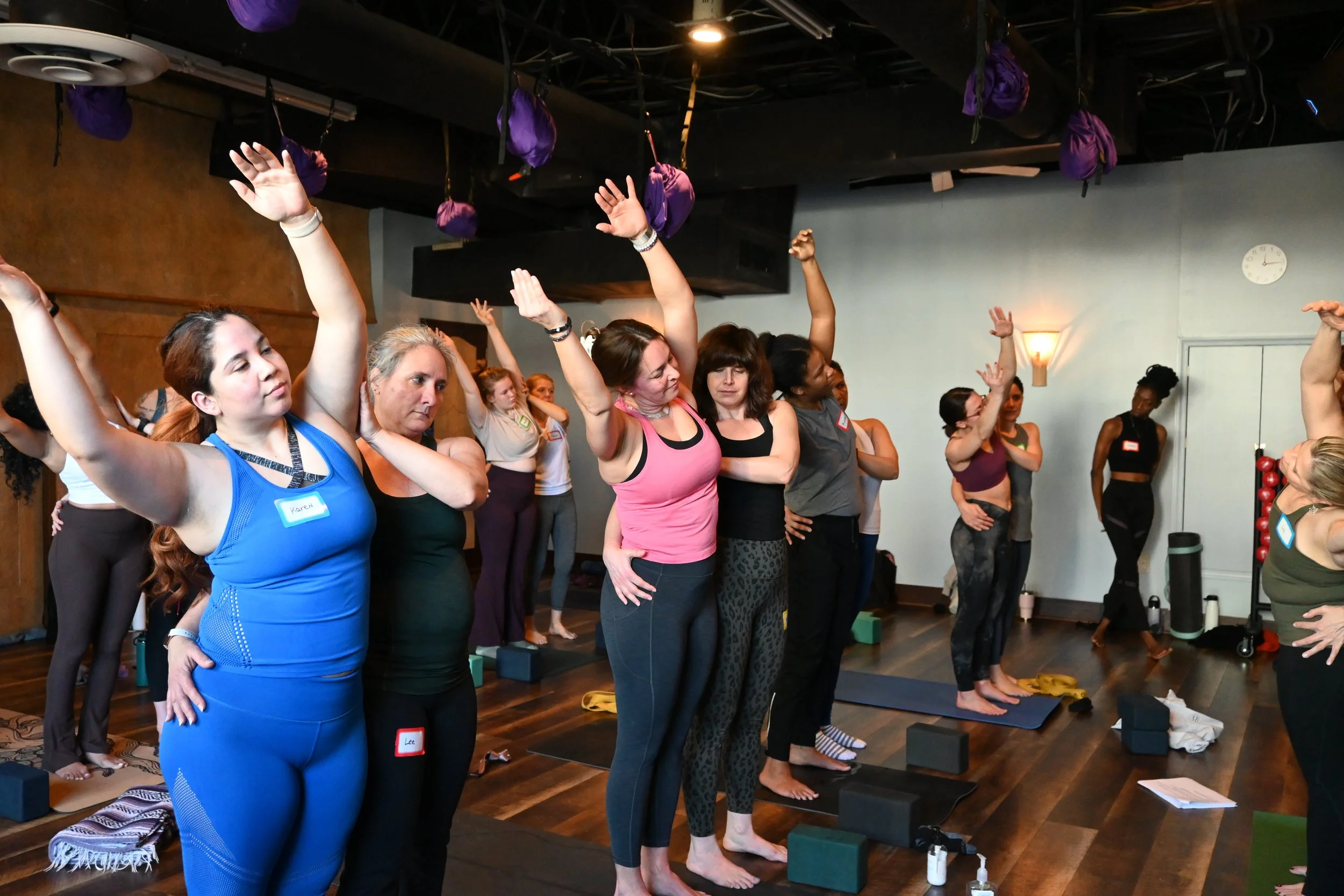 Group of women participating in a yoga or stretching class, standing on mats and blocks with arms raised and relaxed in a room with a wooden floor and purple hanging bags from the ceiling.