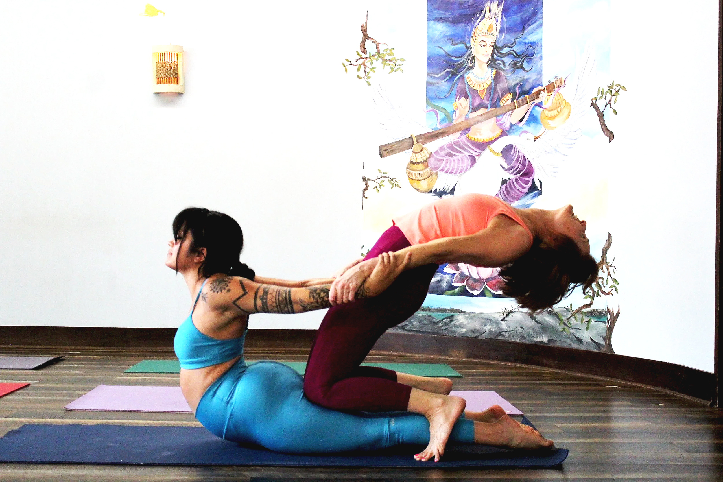 Two women practicing partner yoga in front of a colorful mural of a goddess on a white wall. One woman with tattoos is in a backbend pose, supporting the other woman leaning backward on her arms and legs.