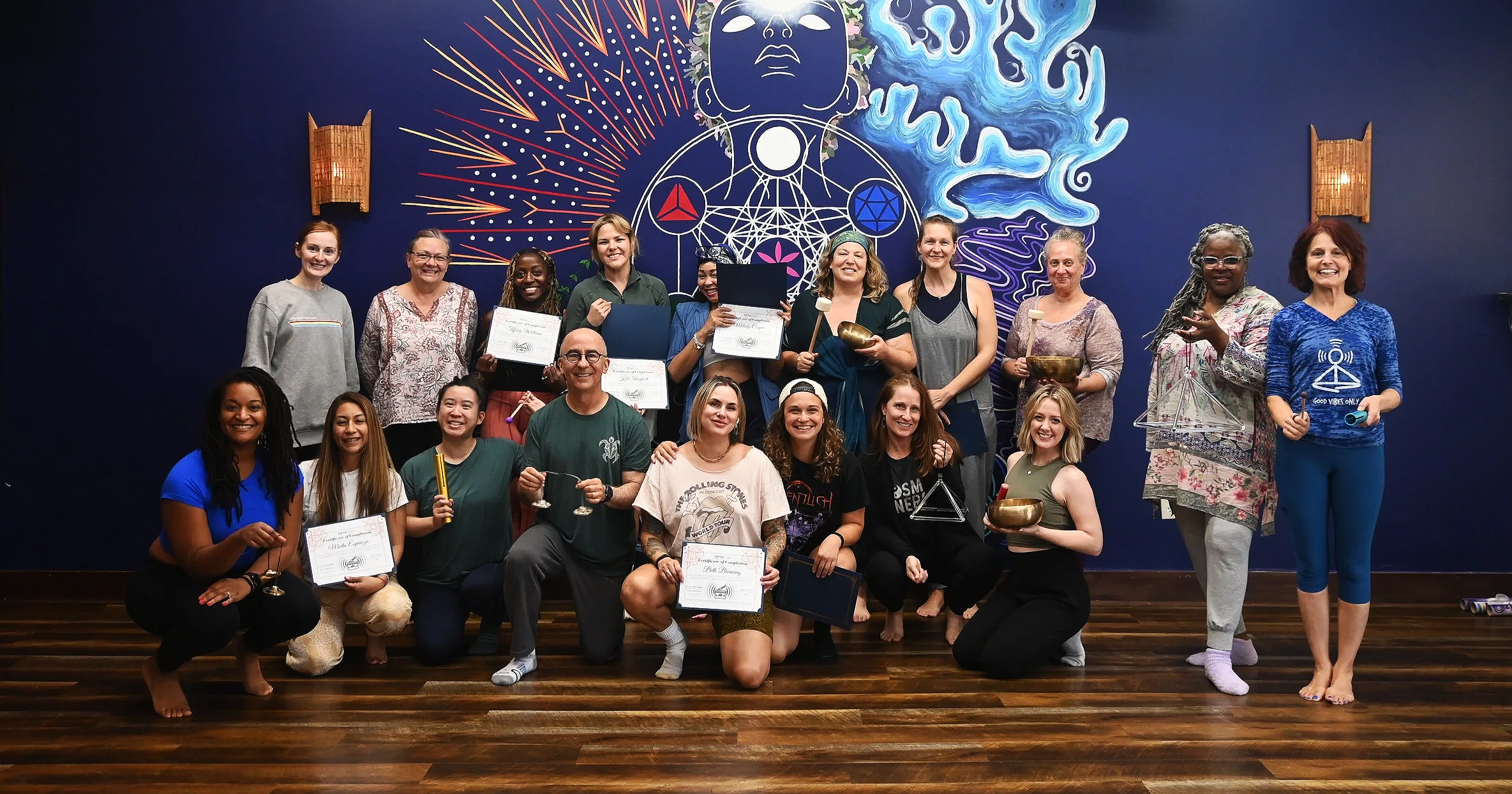 Group of women and men posing together with certificates and singing bowls in front of a colorful mural, some kneeling and some standing on a wooden floor.