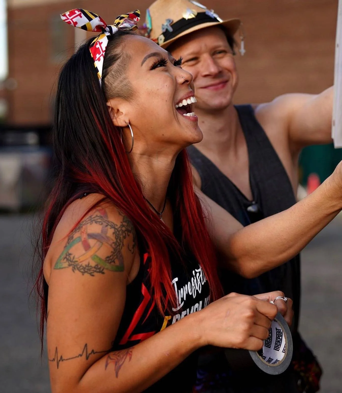 A woman with red hair, tattoos, and hoop earrings smiling and holding a medal, with a man in the background taking a selfie. They are outdoors, and the woman is wearing a headband with a red, black, yellow, and white pattern.