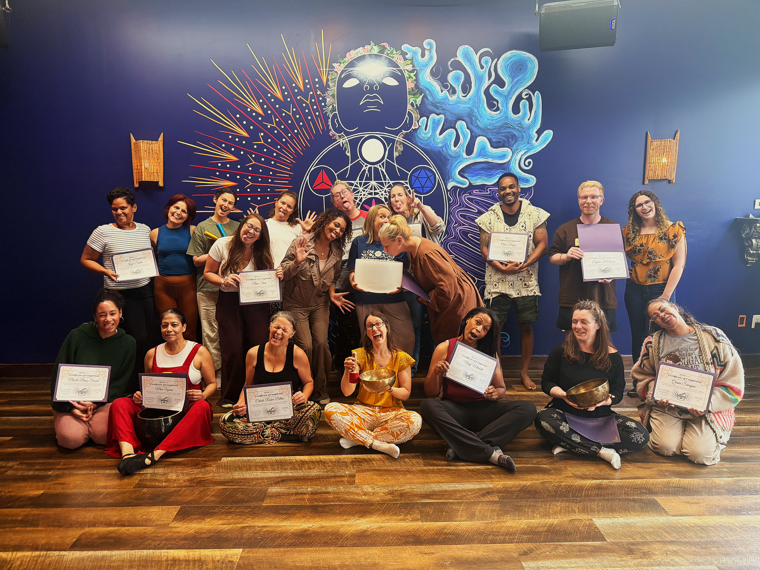 A group of people on a stage holding certificates and surprise awards, posing for a photo. The backdrop features a colorful mural with abstract figures, a face, and decorative designs.