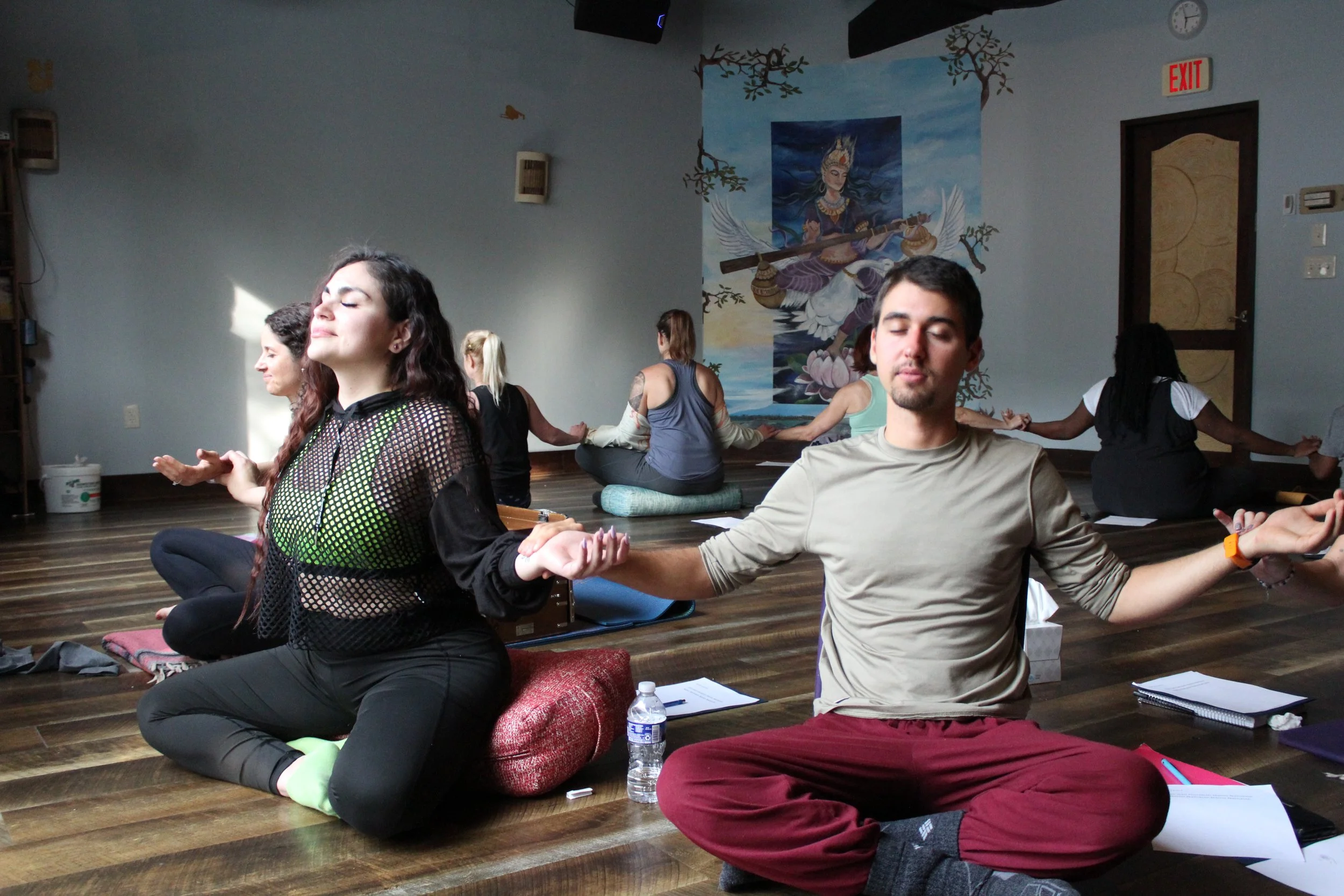 People meditating in a yoga class, sitting cross-legged on mats, with eyes closed and hands resting on knees, in a room with a large mural of a woman playing a musical instrument.