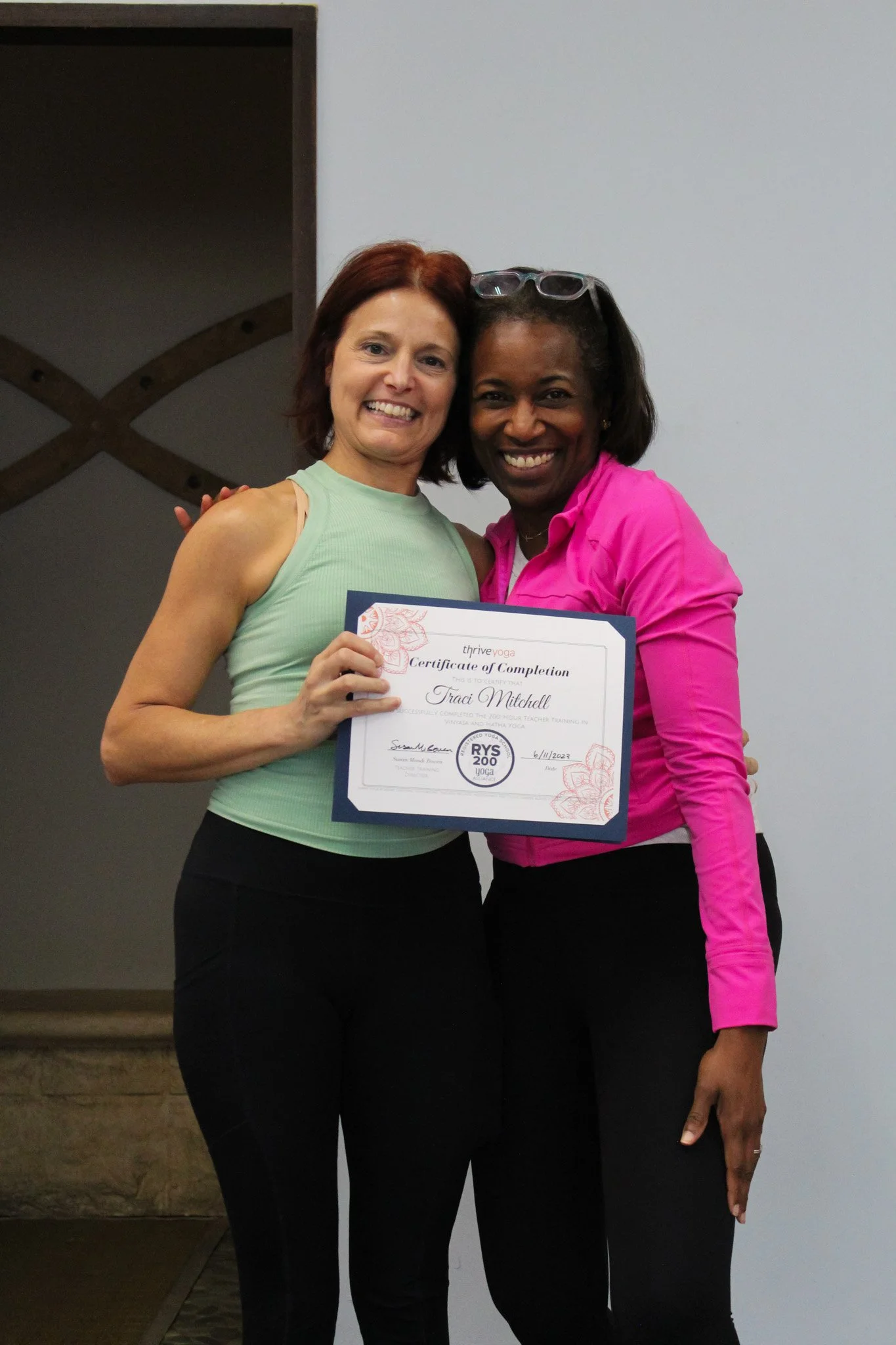 Two women standing close together, smiling, one holding a certificate of completion, against a plain wall background.