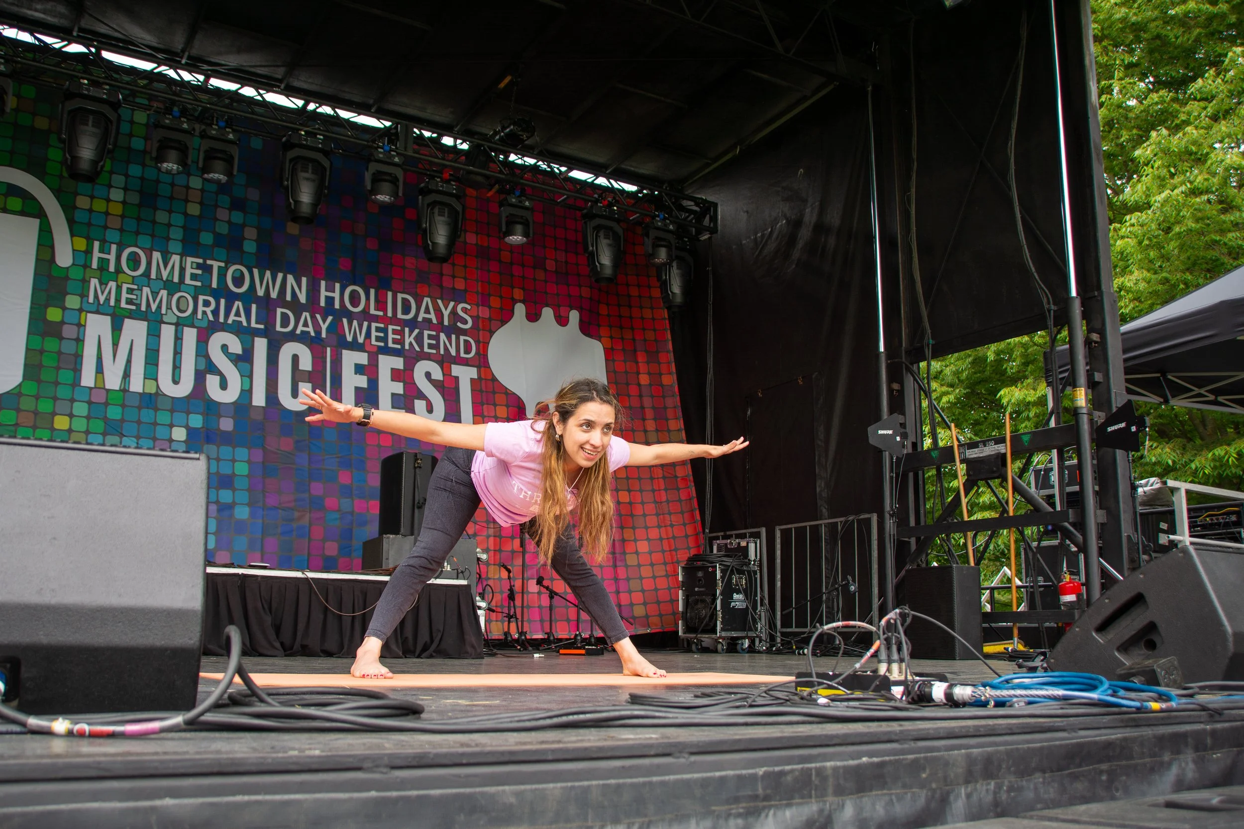 A young woman practicing yoga on stage during an outdoor music festival with a colorful backdrop and stage lighting, surrounded by sound equipment and cables.
