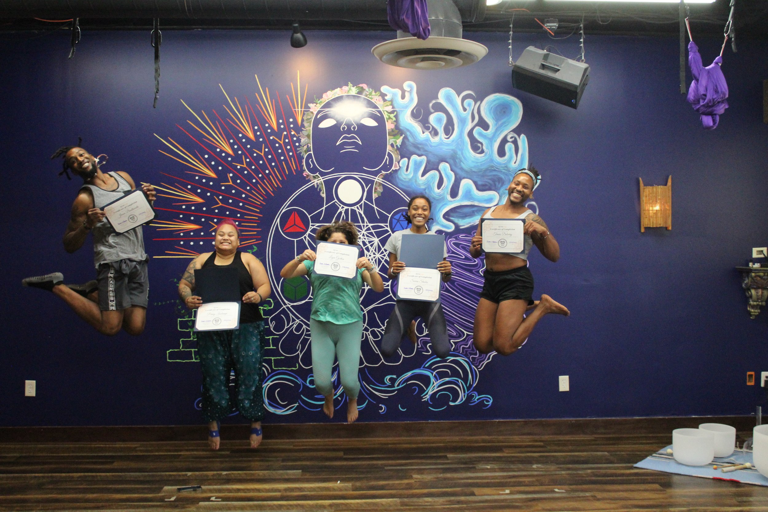 Six people jumping in front of a colorful mural with a stylized face and abstract designs. Four women and two men, all smiling and holding certificates, in a fitness or yoga studio.
