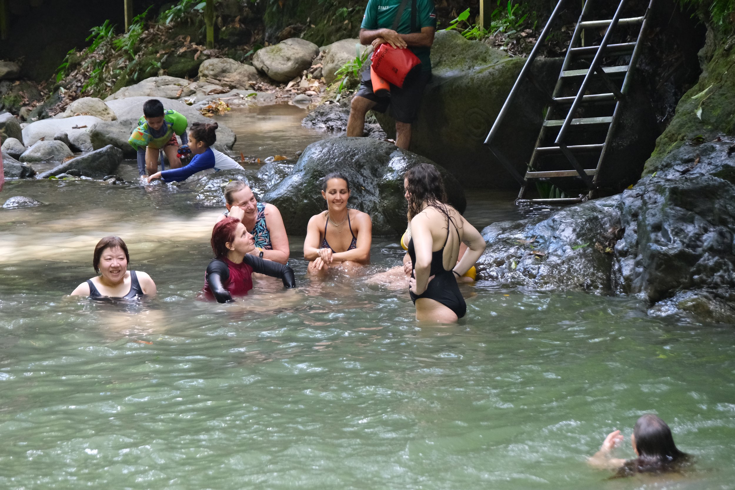 People swimming and relaxing in a natural stream in a jungle setting, with rocks, greenery, and a metal ladder on the side.