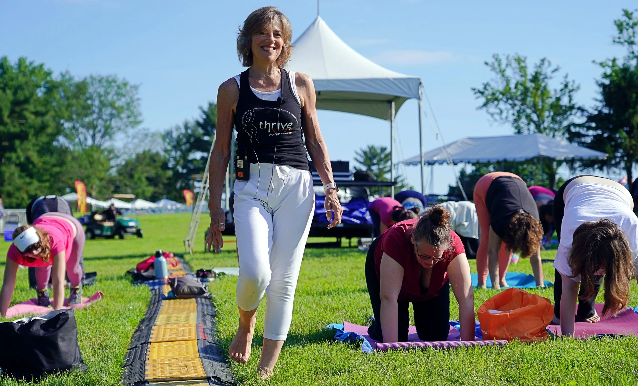 A woman walking barefoot on a path during a yoga session outdoors, with other participants in yoga poses on mats on the grass and a white canopy in the background on a sunny day.