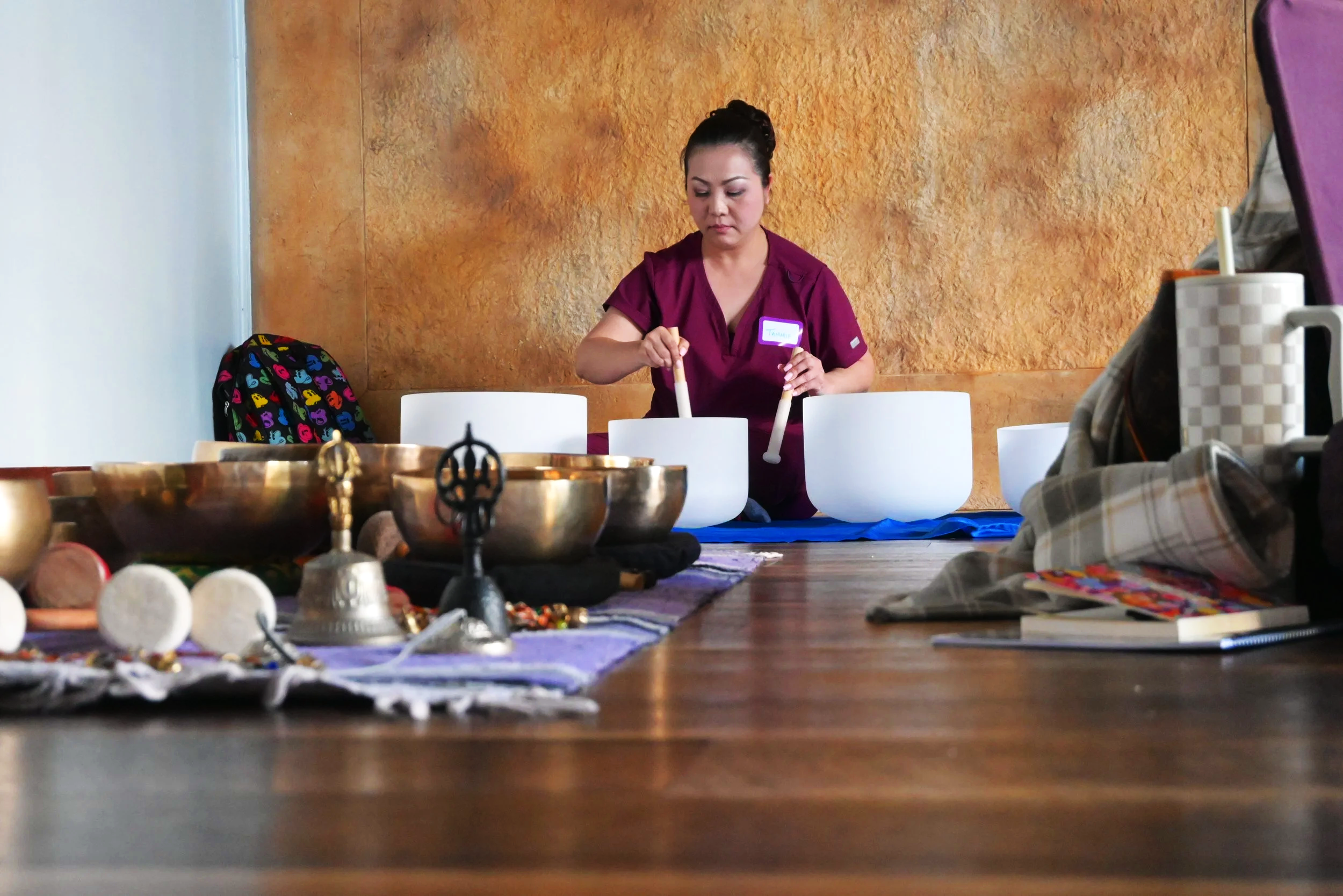 A woman with dark hair tied up, wearing a purple uniform, appears to be meditating or engaging in a spiritual practice indoors, with a textured golden-brown wall behind her. In the foreground, a table displays various metallic and decorative objects, possibly related to spiritual or cultural rituals.