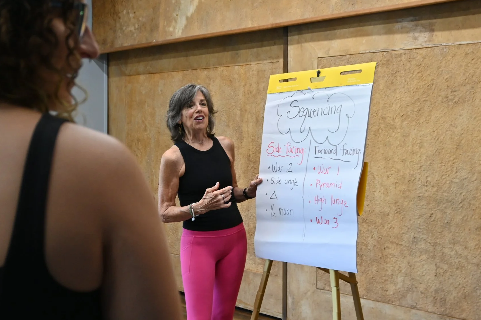 A woman in a black tank top and pink pants giving a presentation about sequencing, with a large sheet of paper on a stand behind her displaying key points about side facing and forward facing sequencing techniques listed in red and black ink.
