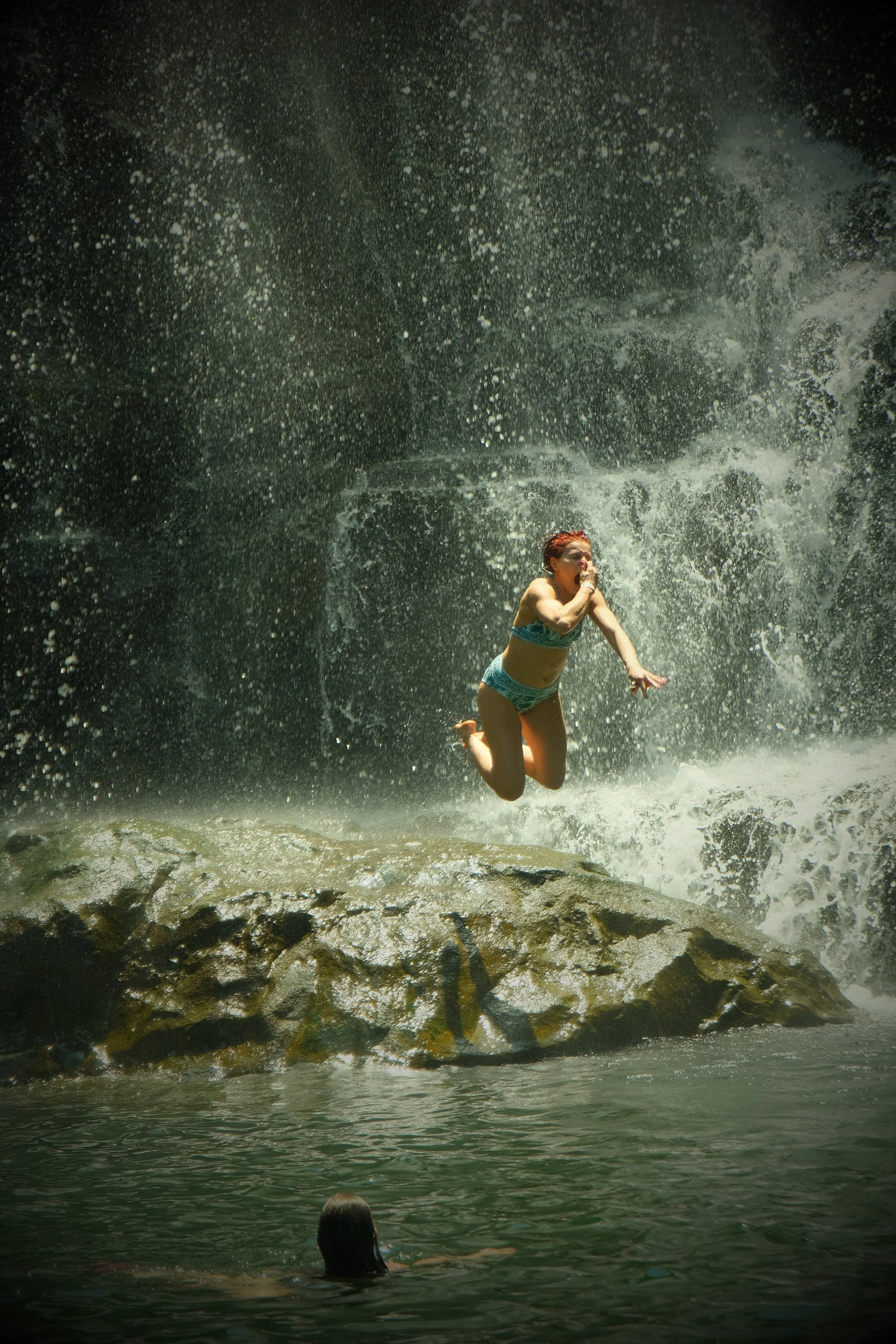 A woman in a bikini jumping off rocks into the water at a waterfall, with another person in the water watching.