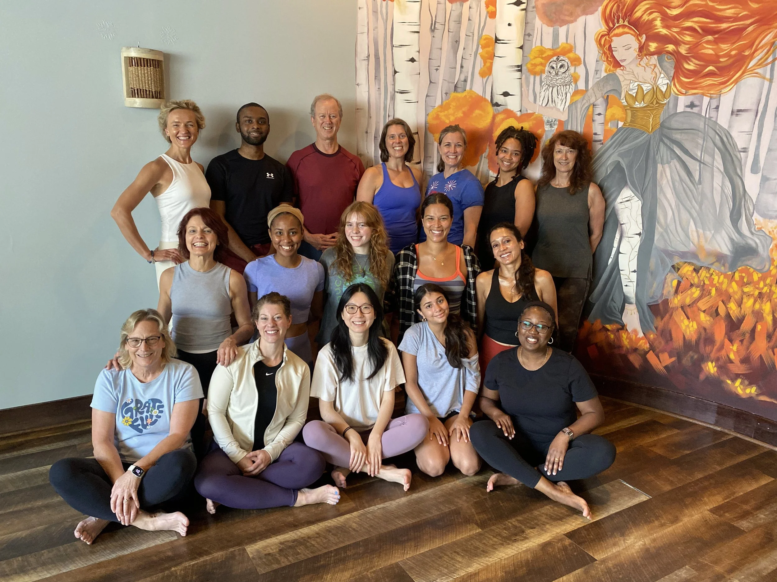 A group of diverse women and a man posing for a photo in a room with a mural of a woman with flowing red hair, an owl, and autumn trees in the background. Some are sitting on the hardwood floor, others are standing behind them, all smiling.