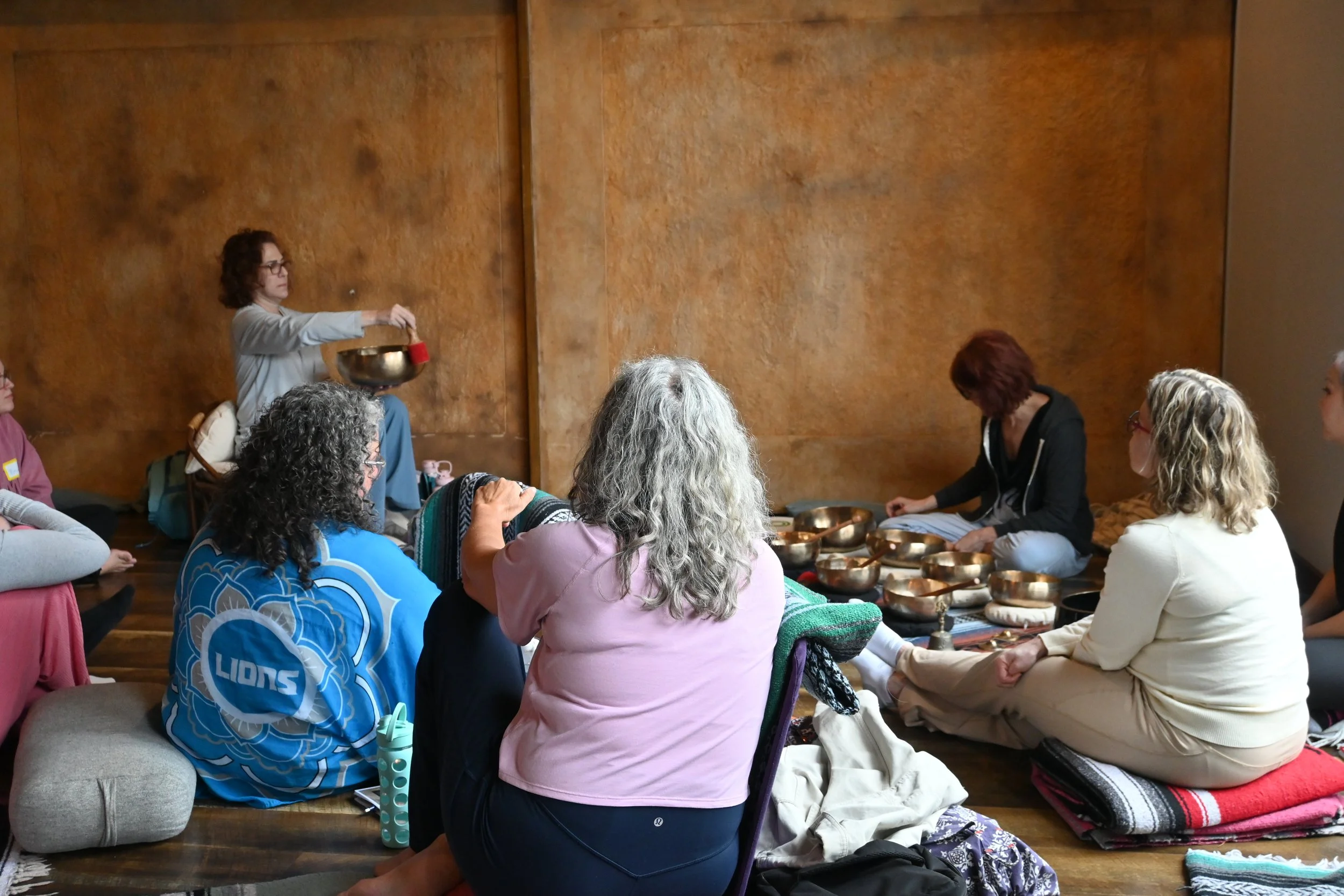 A group of women sitting on the floor in a circle, participating in a meditation or sound healing session with singing bowls. One woman is playing a singing bowl while others are seated around her, some with eyes closed and others looking attentively
