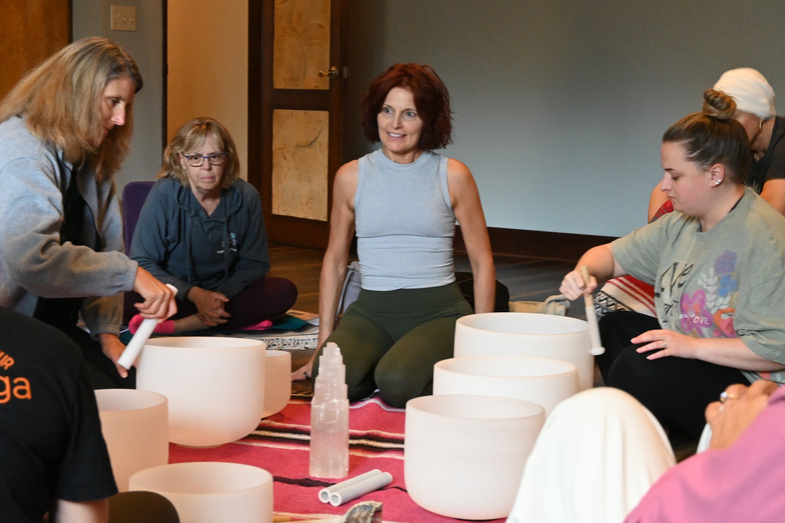 A group of women sitting on the floor around large white singing bowls, with some playing mats and tubes, in an indoor room with wooden door and wall paneling.