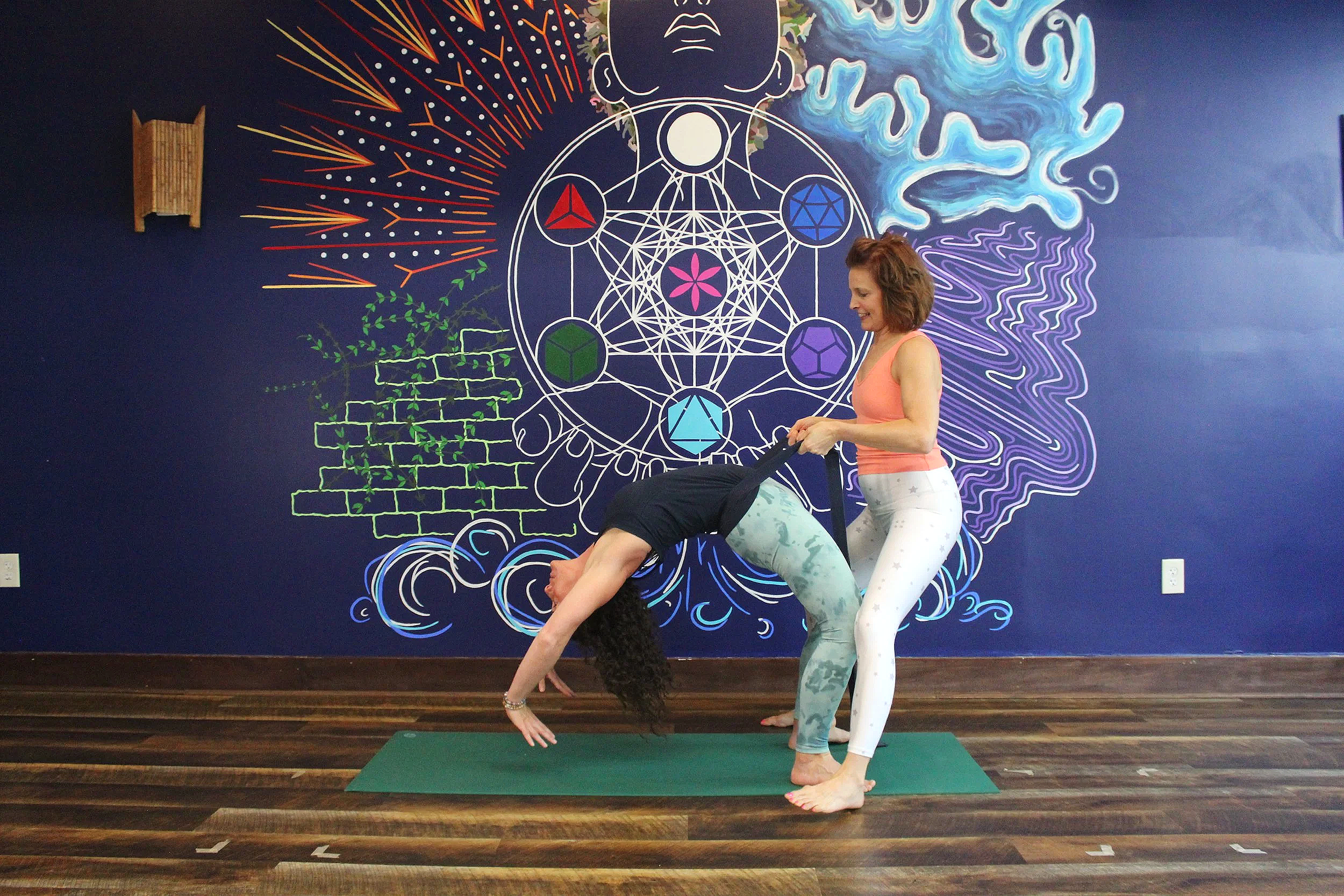 Two women practicing yoga indoors, one in a downward dog pose on a green yoga mat and the other standing beside her holding her ankles, with a vibrant mural featuring geometric patterns and colorful designs on the dark blue wall in the background.