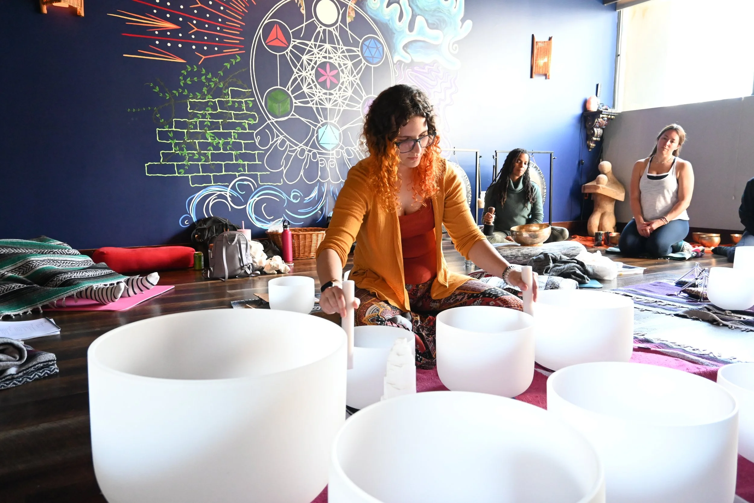 Women participating in a sound healing session with crystal singing bowls in a room decorated with neon art and a large blue wall.