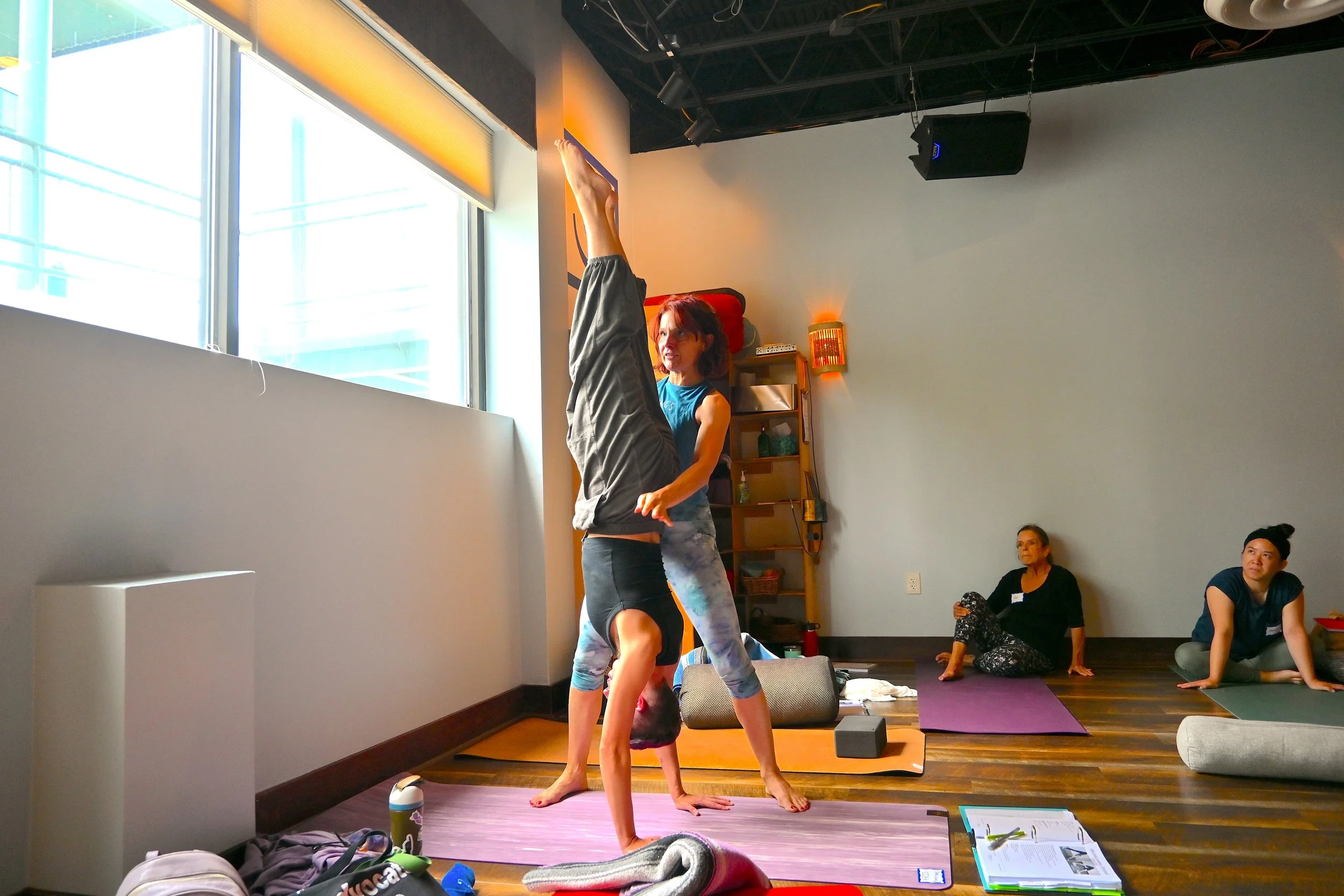 A group of people practicing yoga in a studio, focusing on an instructor demonstrating an acro yoga pose with a student on their hands and feet. Participants are on yoga mats, watching the instructor, with large windows on the side providing natural 