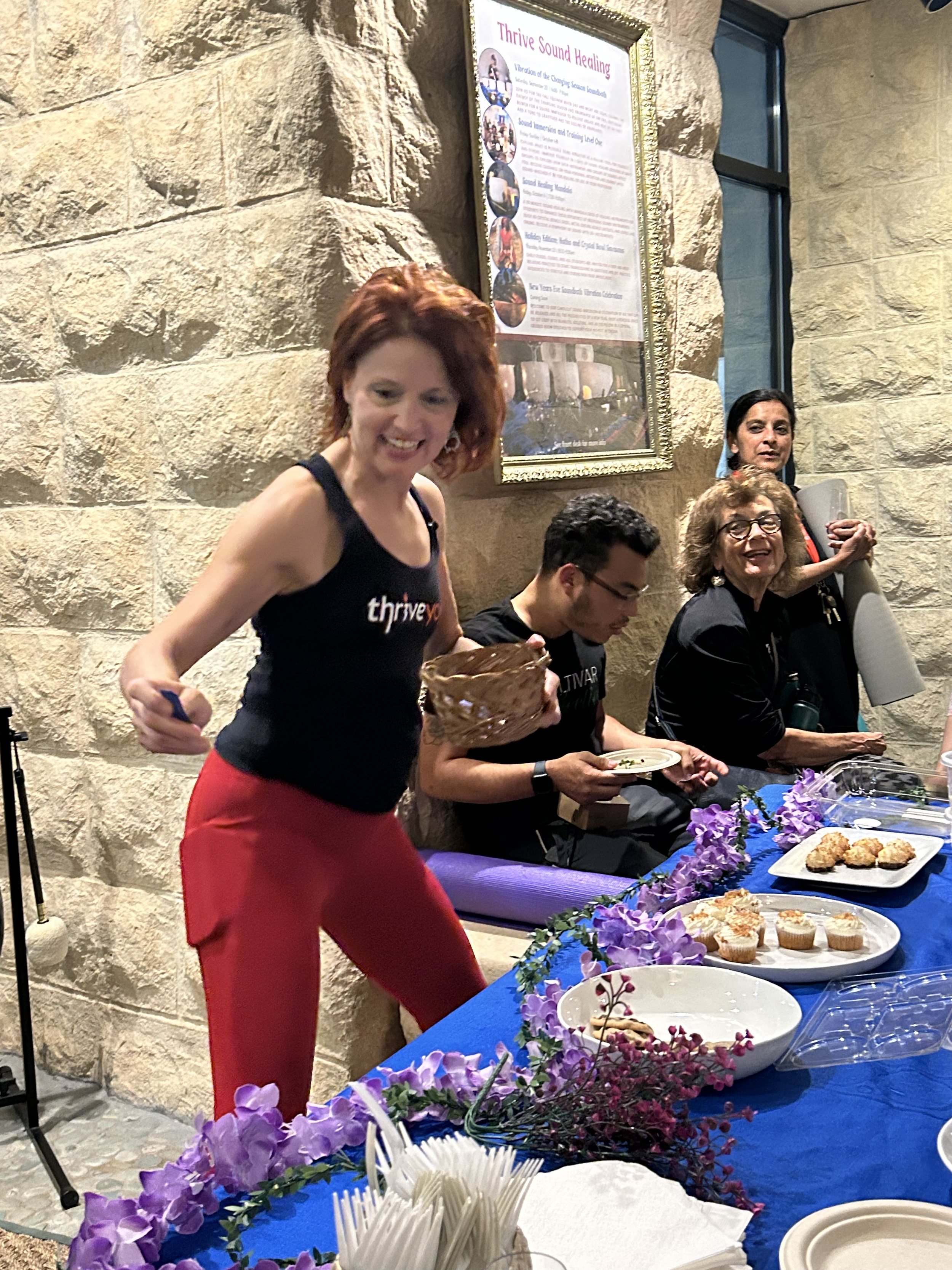 A group of people at a table with food, decorated with purple flowers, in front of a stone wall with a framed poster titled 'Thrive Sound Healing'. A woman with red hair wearing a black tank top and red pants is smiling at the camera, holding a wooden bowl. Two women and two men are seated at the table, with one woman holding a roll of paper and the man eating from a plate.