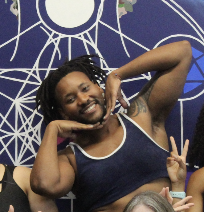 A smiling man with dreadlocks in a tank top, making a playful pose with his hands on his face, standing in front of a blue wall with white geometric and circular designs.