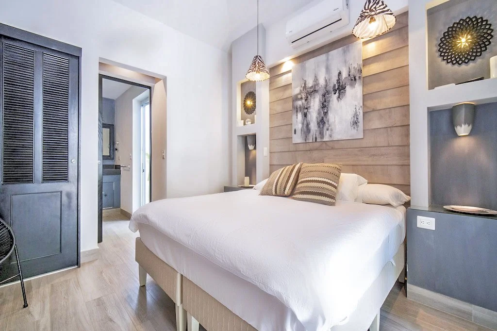 A modern bedroom with a large white bed, decorative pillows, wooden accent wall, abstract art, and built-in wall niches with lighting. There is a door leading to a bathroom and curtains with zebra print on bedside lamps.