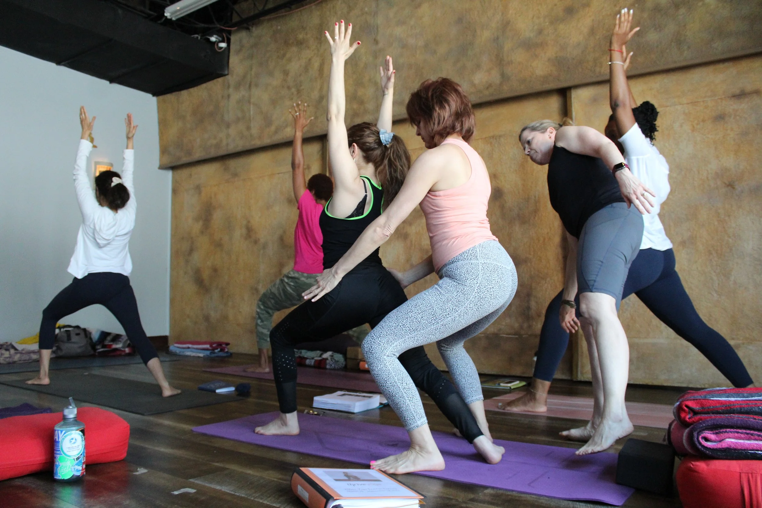 Group of people participating in a yoga class, practicing warrior pose on yoga mats in a studio with a wooden wall.