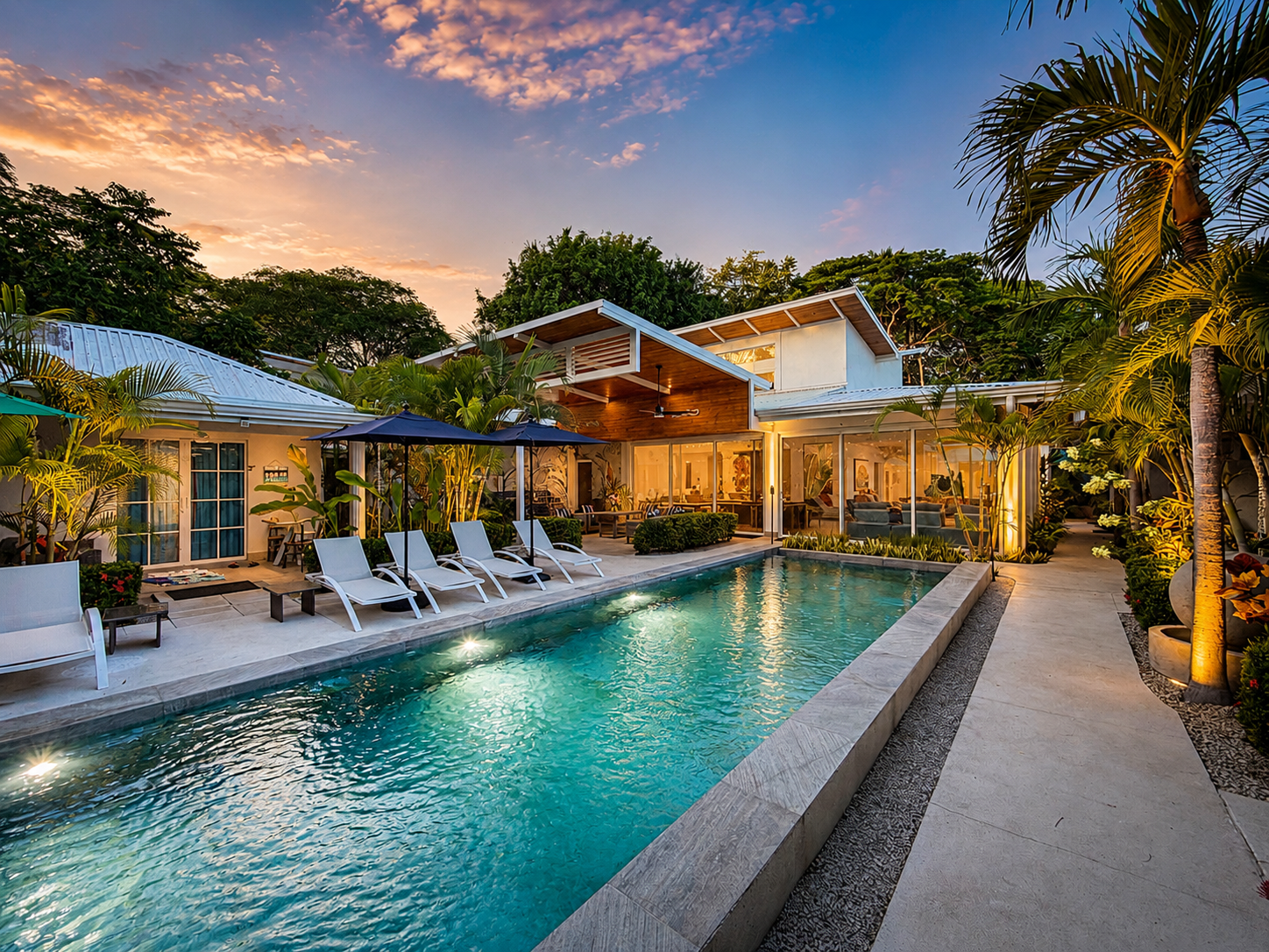 A backyard with a swimming pool surrounded by lounge chairs, umbrellas, lush greenery, and a modern house with large windows at sunset.