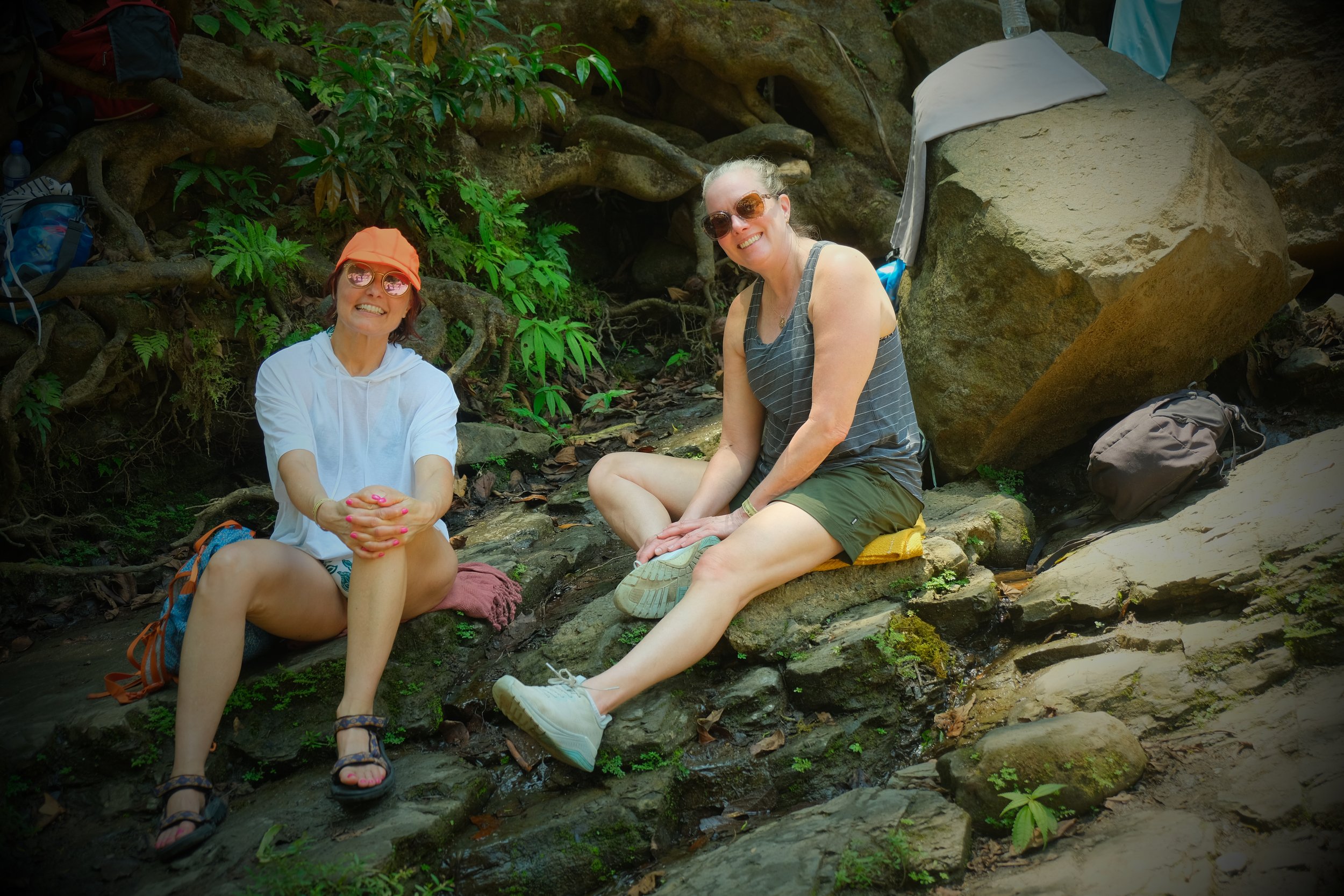 Two women sitting by a small waterfall in a lush, green forest. They are smiling and wearing sunglasses; one is in an orange cap and white hoodie, the other in a striped tank top and green shorts. They have backpacks and hiking gear around them.
