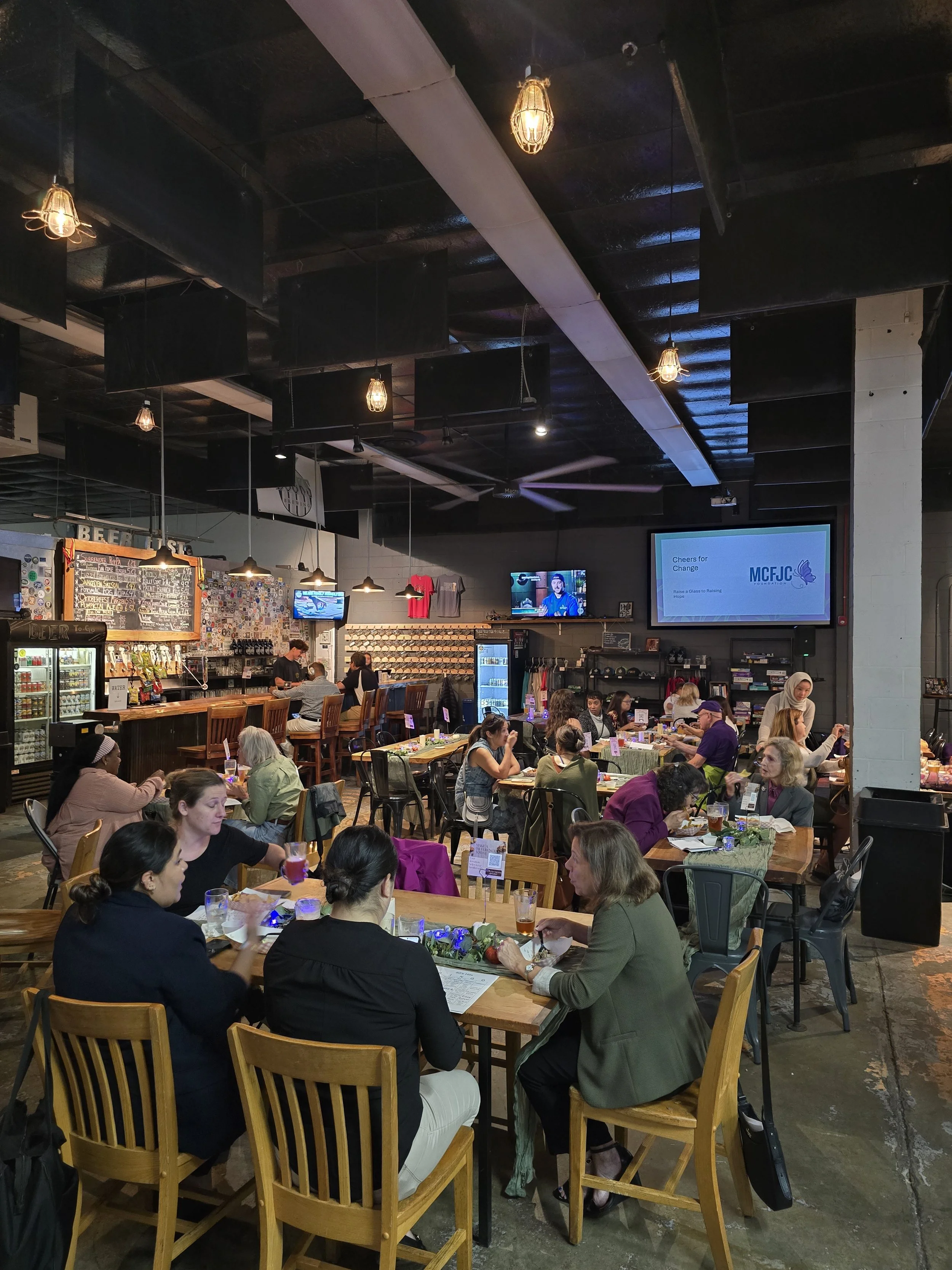 People sitting at tables in a busy restaurant or cafe, engaging in conversations and enjoying meals, with warm lighting and a modern industrial decor.