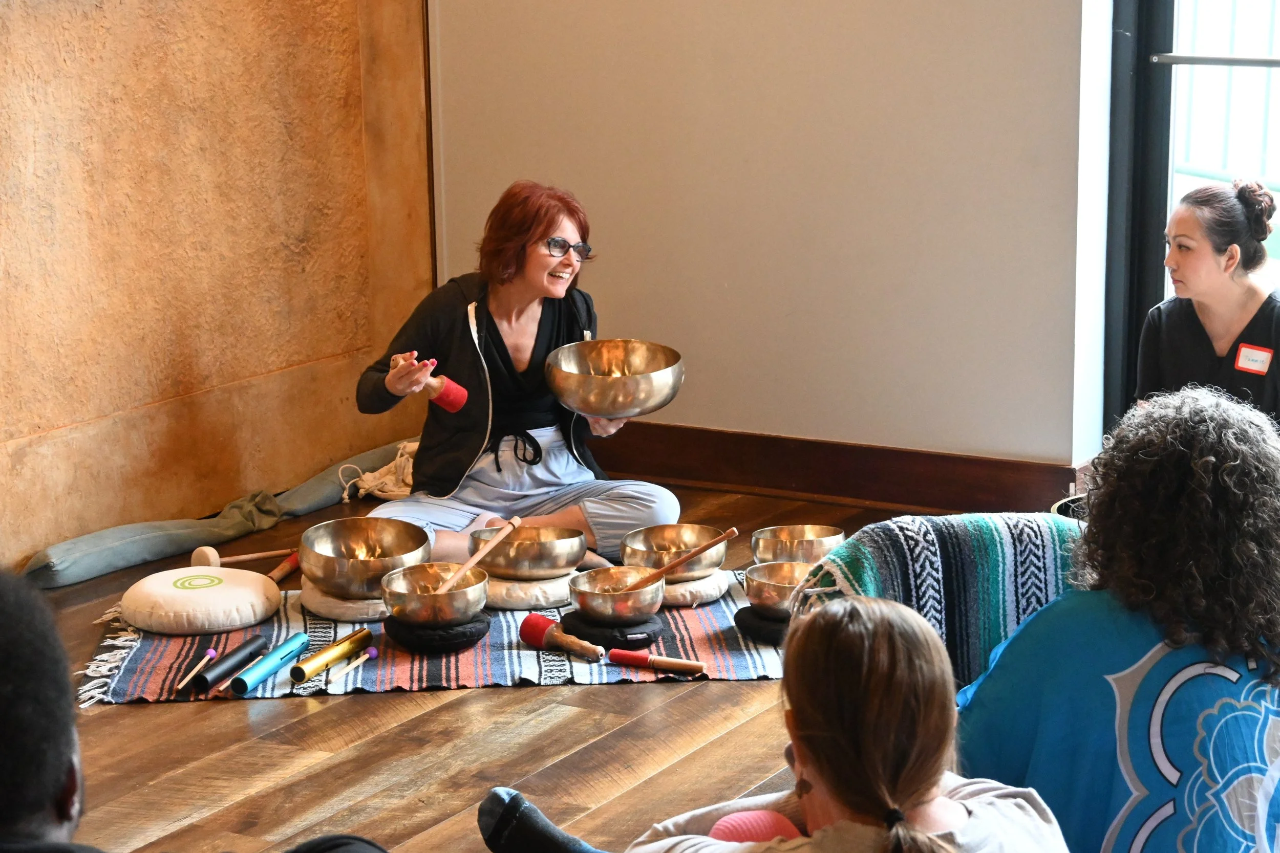 A woman with glasses and short red hair sitting on a cushion, engaging with a group of children during a music session with singing bowls and recorders in a room with wooden floors and beige walls.