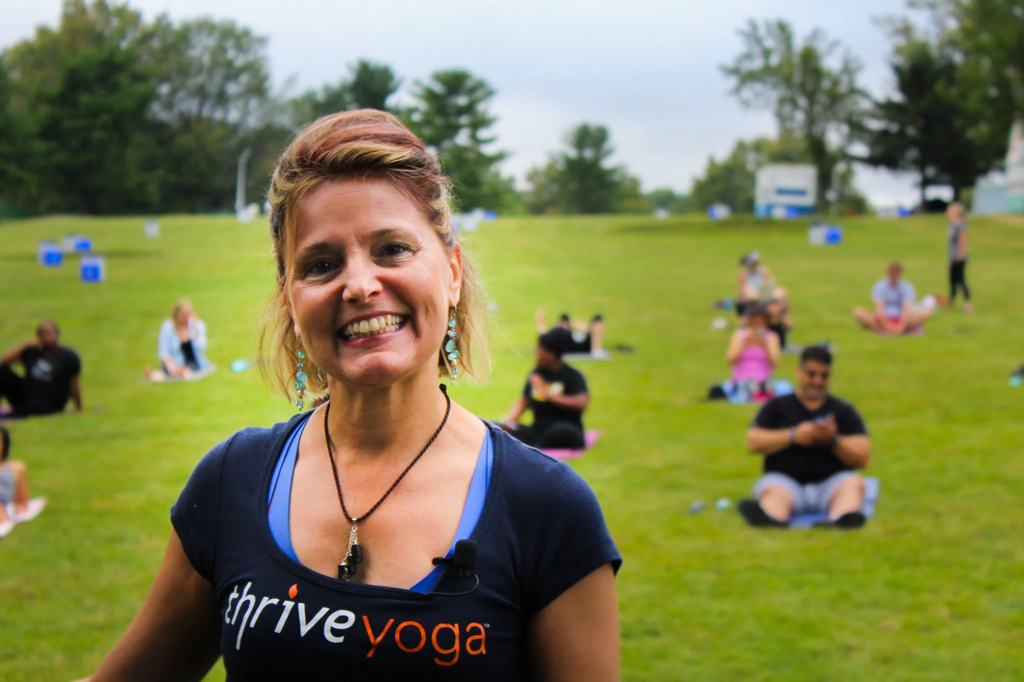 Smiling woman with short red hair, wearing a blue t-shirt with 'thrive yoga' written on it, standing outdoors in a park with people practicing yoga in the background.