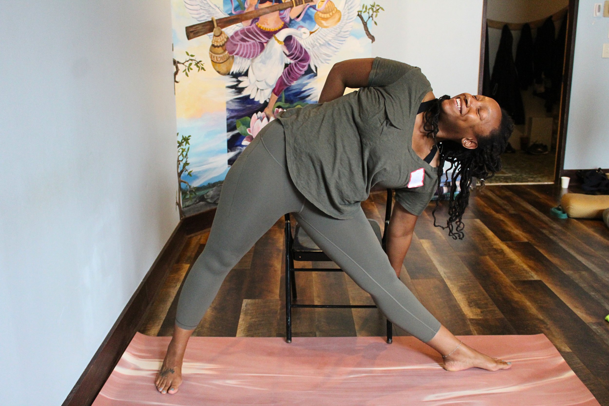 A woman with curly hair smiling and doing a side stretch pose on a yoga mat. She is in a room with a colorful mural of a bird in the background.