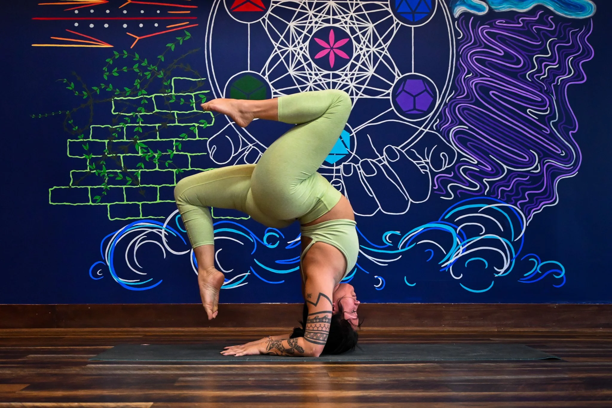 A woman in a yoga pose performing a handstand with her legs bent and feet touching her head, on a dark yoga mat in front of a colorful mural. The mural has intricate white, green, purple, and blue designs, including a tree, geometric patterns, and flowing lines.