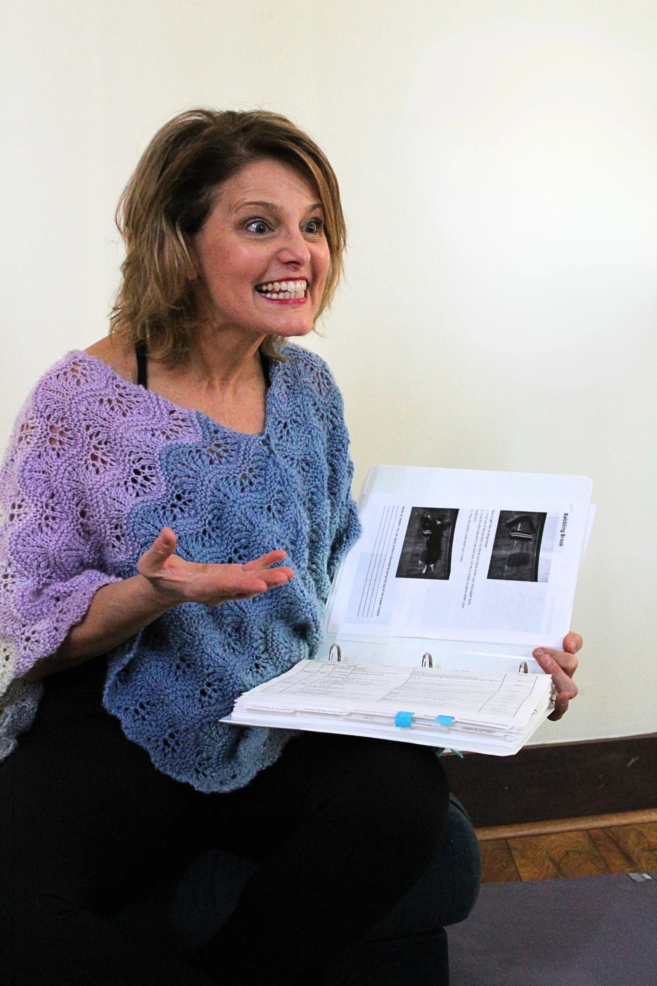 A woman with short, wavy brown hair, wearing a purple crocheted top, is sitting and holding an open binder with documents. She appears to be speaking and gesturing with her left hand, with a white wall and wooden floor in the background.