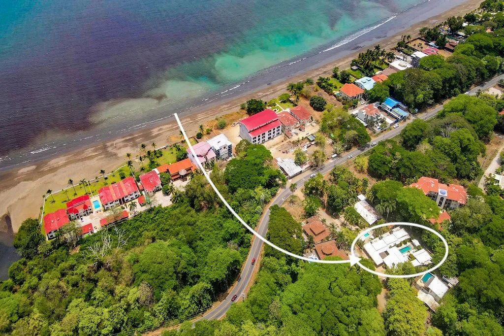 Aerial view of a coastal neighborhood with houses, trees, and a beach, showing a white arrow indicating a round trip from a house near the road to the beach.