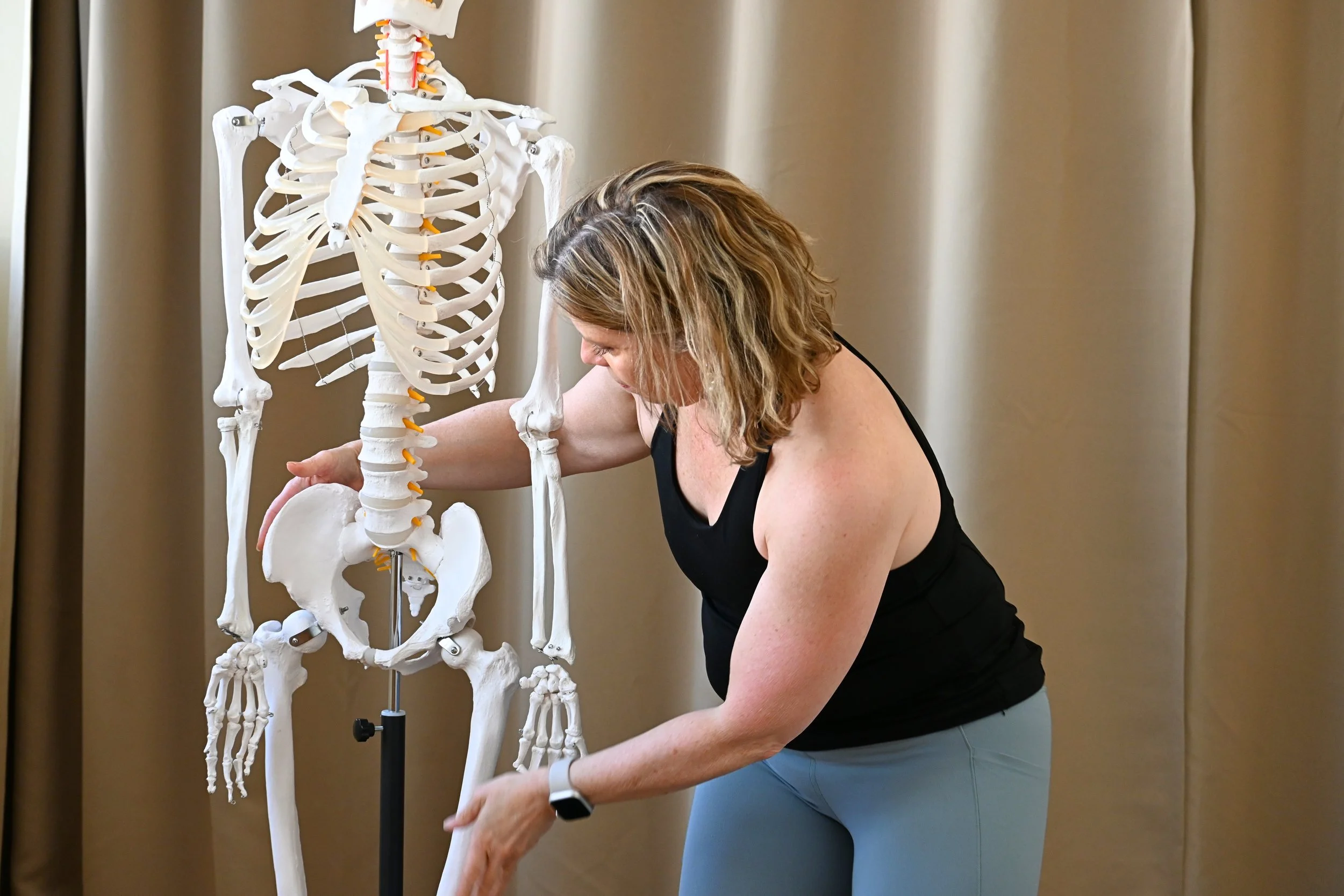 A woman leaning over and examining a life-size human skeleton model in a room with beige curtains.