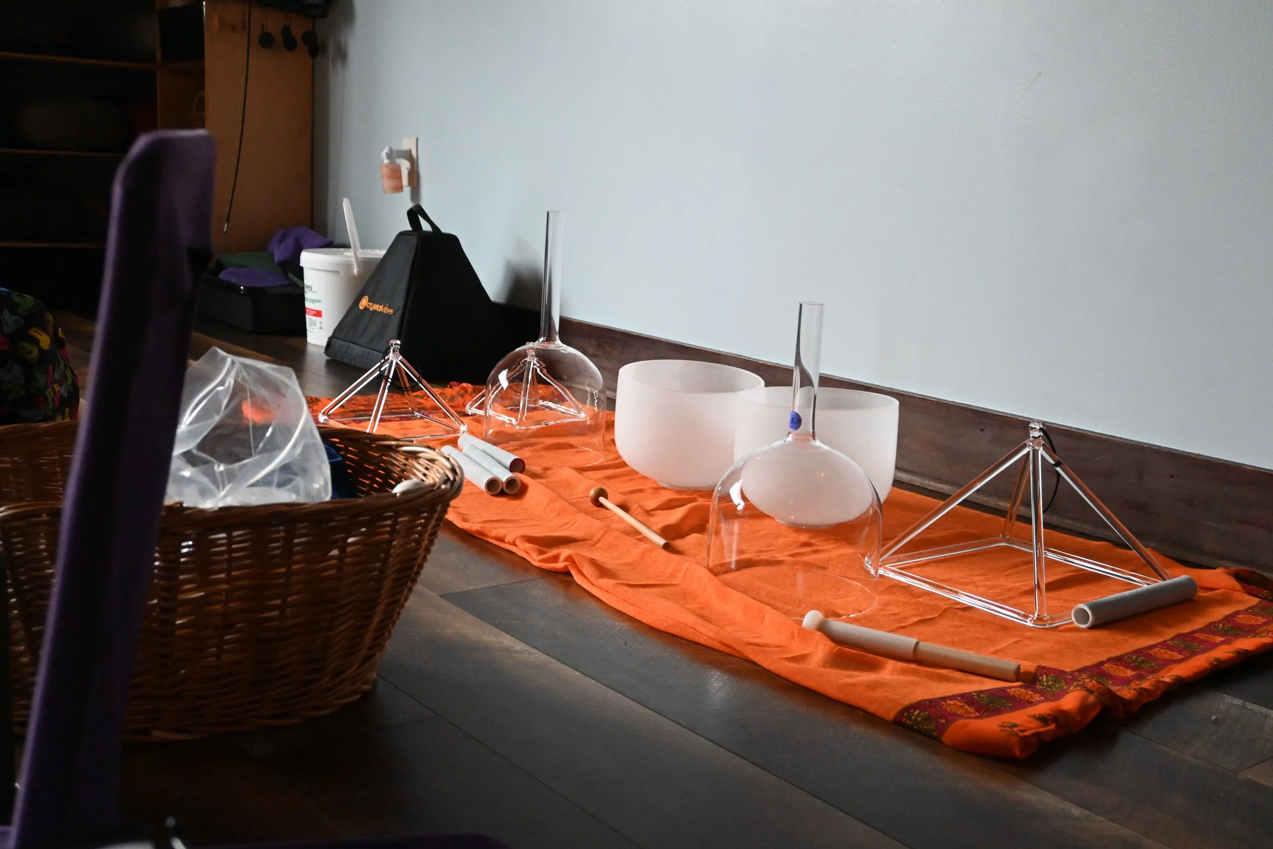 Laboratory setup with two white singing bowls, glass bells, and metal stand, placed on an orange cloth on a wooden floor, against a plain wall with scientific glassware nearby.