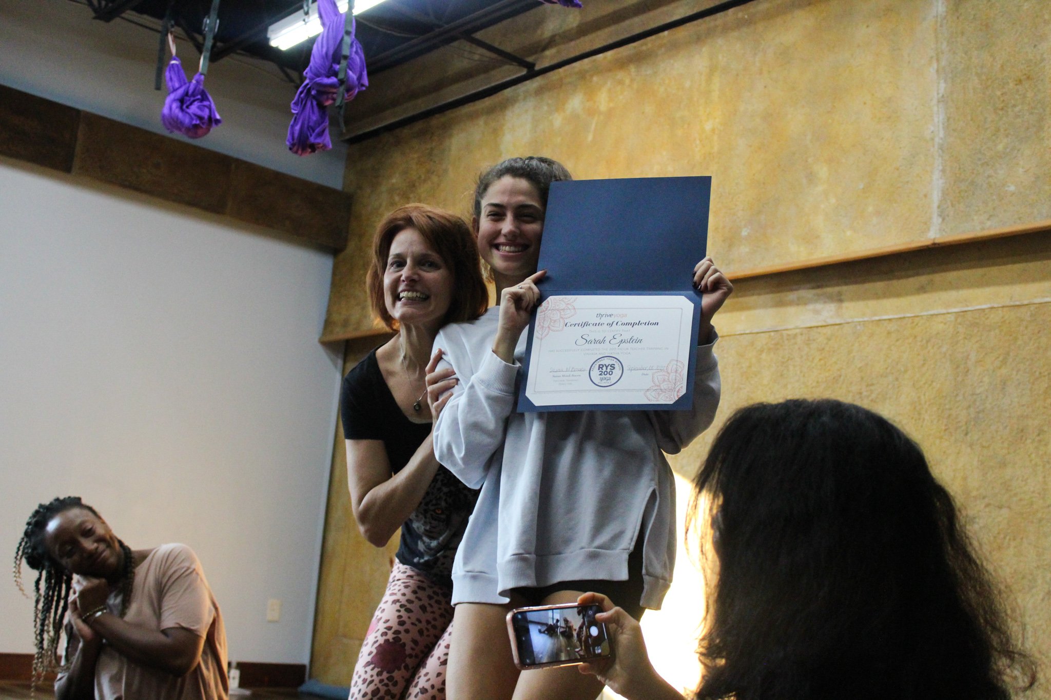 A woman holding a certificate of completion, smiling, with others standing around in a room with a brown textured wall and purple hanging fabric in the background.